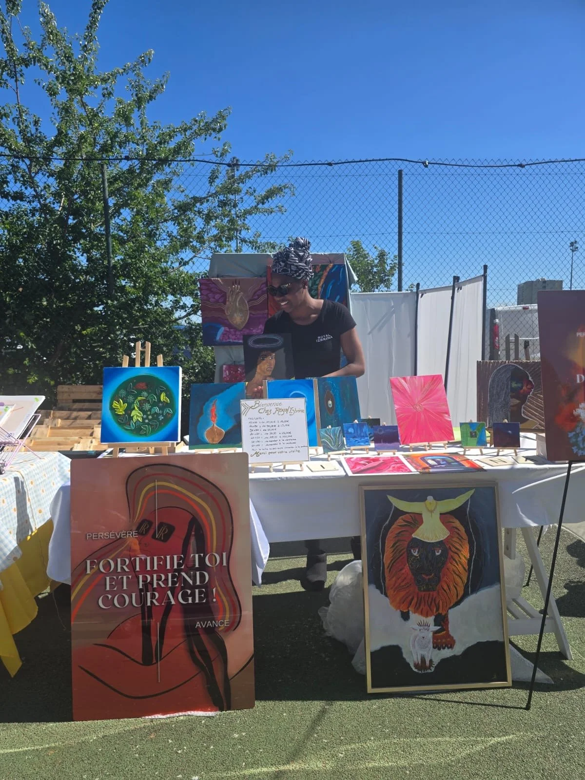 Vente d'œuvres d'art à un stand en plein air sous un ciel bleu, avec une femme portant des lunettes de soleil et un foulard, entourée de peintures colorées représentant des animaux et des motifs abstraits.