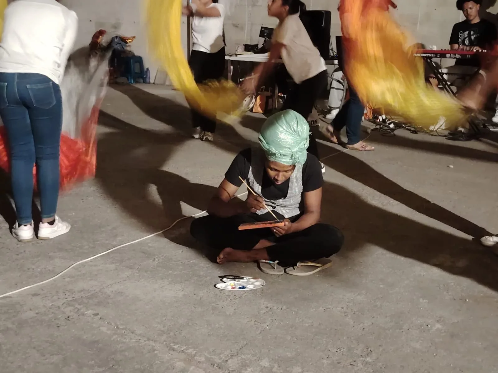 Une femme assise par terre, peignant une toile miniatures, entourée de danseurs avec des voiles colorés lors d'une performance artistique ou culturelle de nuit.