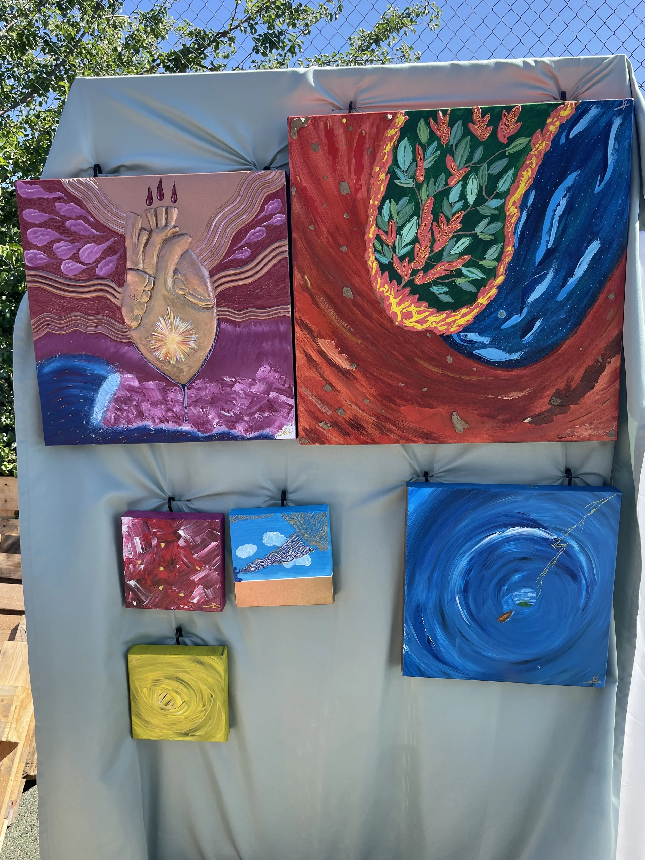 Exposition de cinq peintures abstraites de tailles variées accrochées à un panneau en tissu. La peinture en haut à gauche représente un cœur avec des vagues d'énergie et un éclat lumineux au centre. La peinture en haut à droite montre une spirale de 