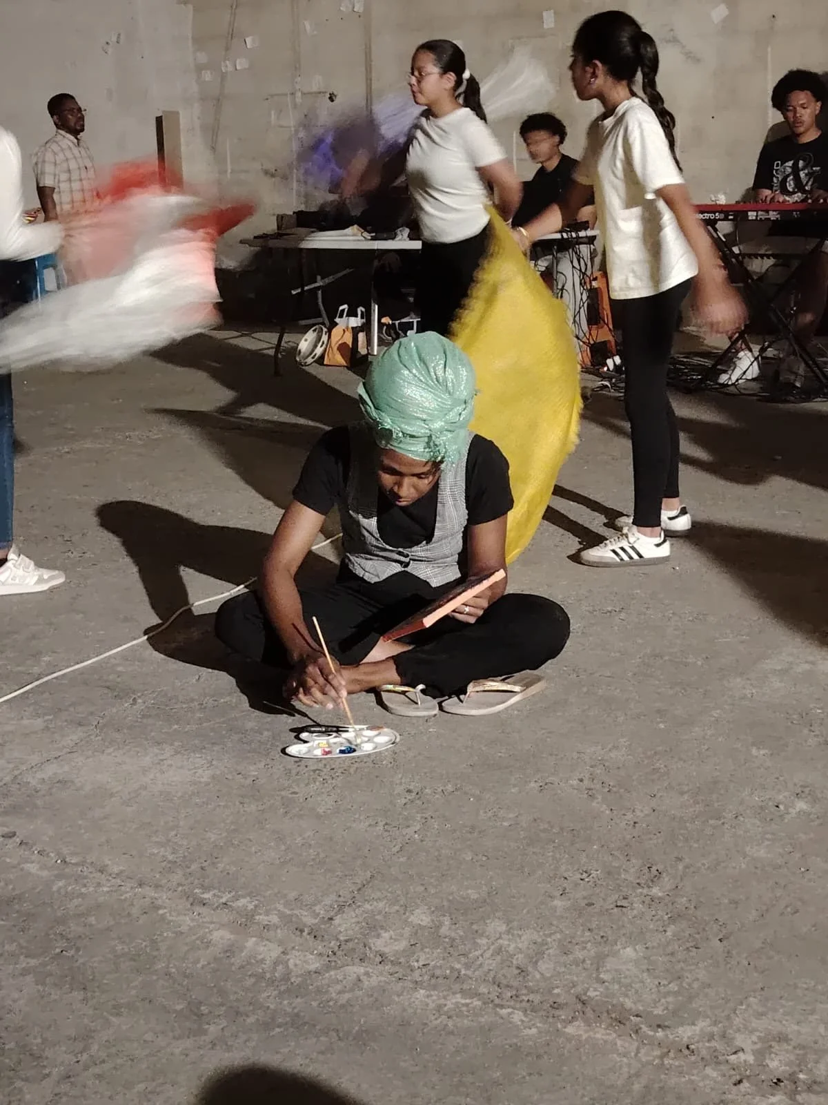 Un groupe de personnes, dont une femme assise par terre et peignant sur une palette avec des pinceaux, semble être dans un espace de performance ou d'art en plein air. D'autres personnes en arrière-plan jouent de la musique et dansent.
