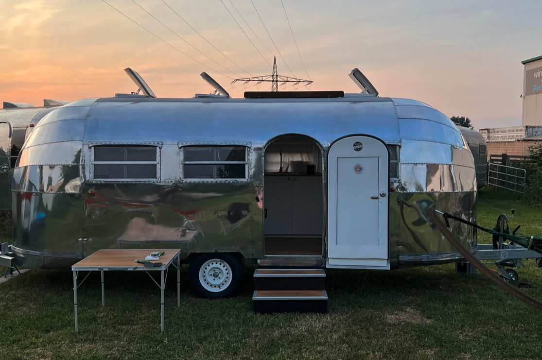 Ein silberner Airstream-Reisewagen vor einem Sonnenuntergang, mit geöffnetem Eingang, einem kleinen Steg, einem Tisch im Vordergrund und einem Fahrrad seitlich.