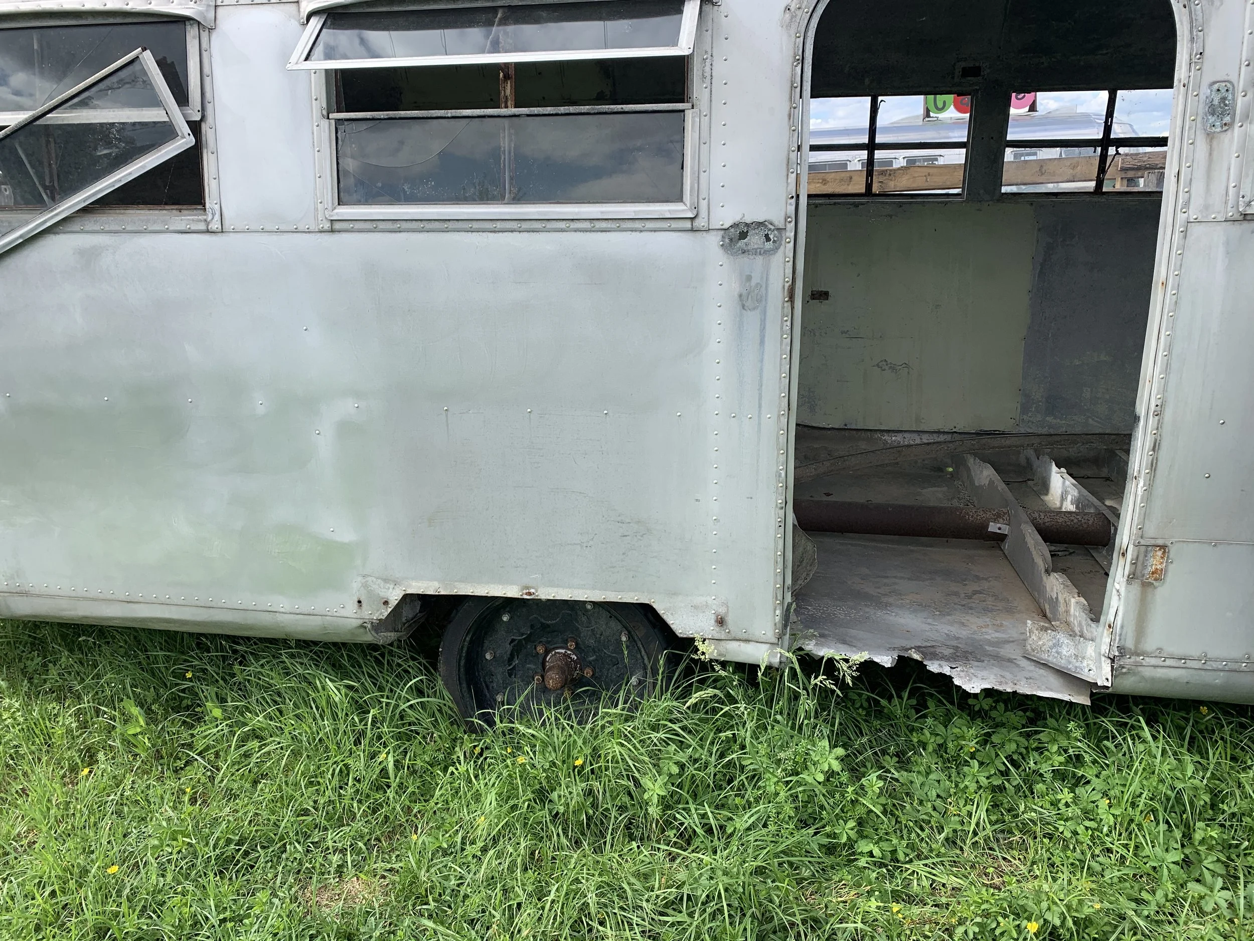 Alte, verlassene Metallreisemobil auf Gras, mit heruntergefallenem Radkasten und offenem Fenster.