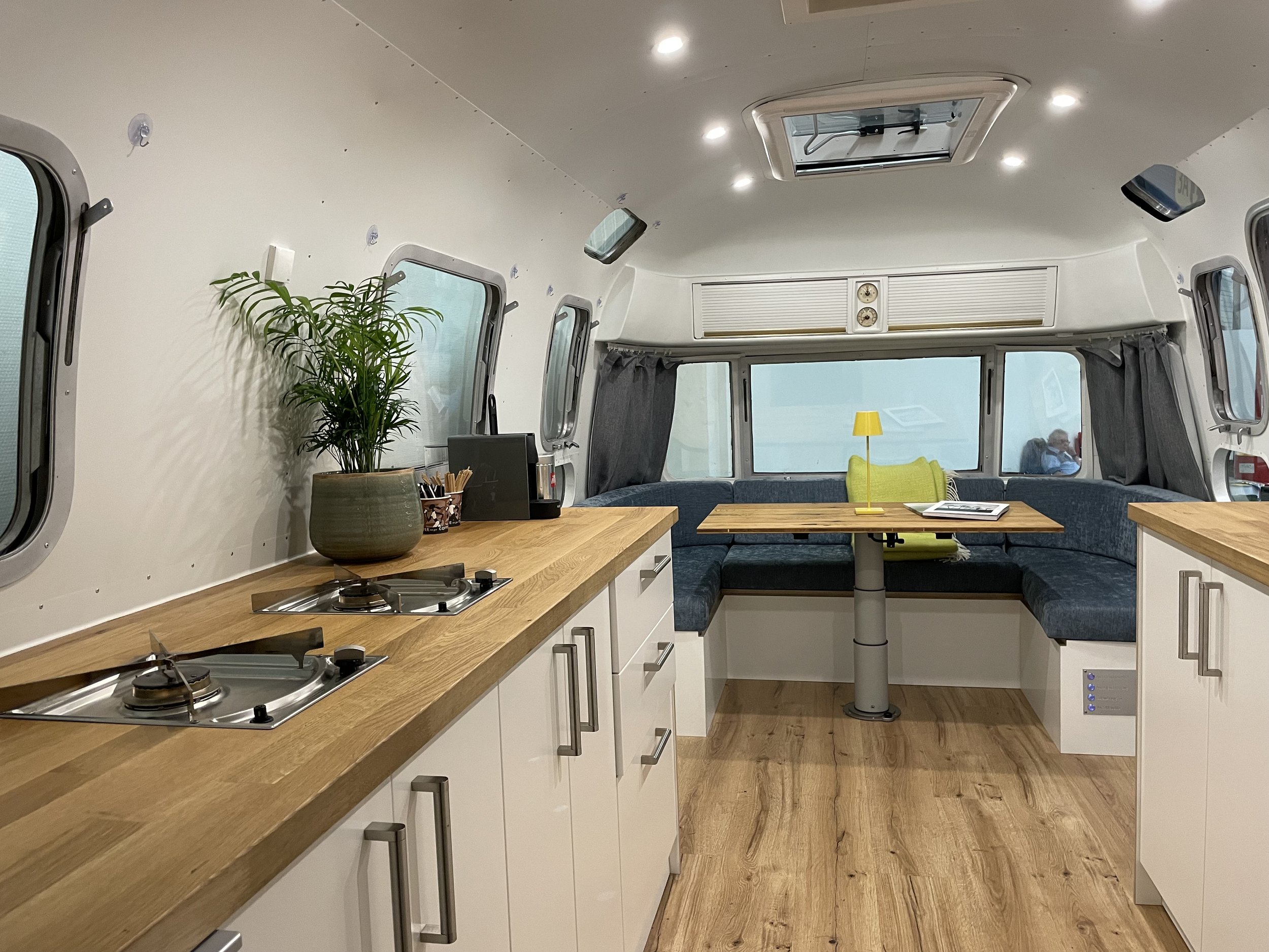 Innenraum einer Wohnmobil-Küche mit Holzarbeitsplatten, weißen Schränken, einer Küchenlampe, einem Pflanzenkübel, einem kleinen Fernseher, einer Sitzbank mit grünem Kissen, einem Tisch, einem Fenster mit grauen Vorhängen und einem Dachfenster.