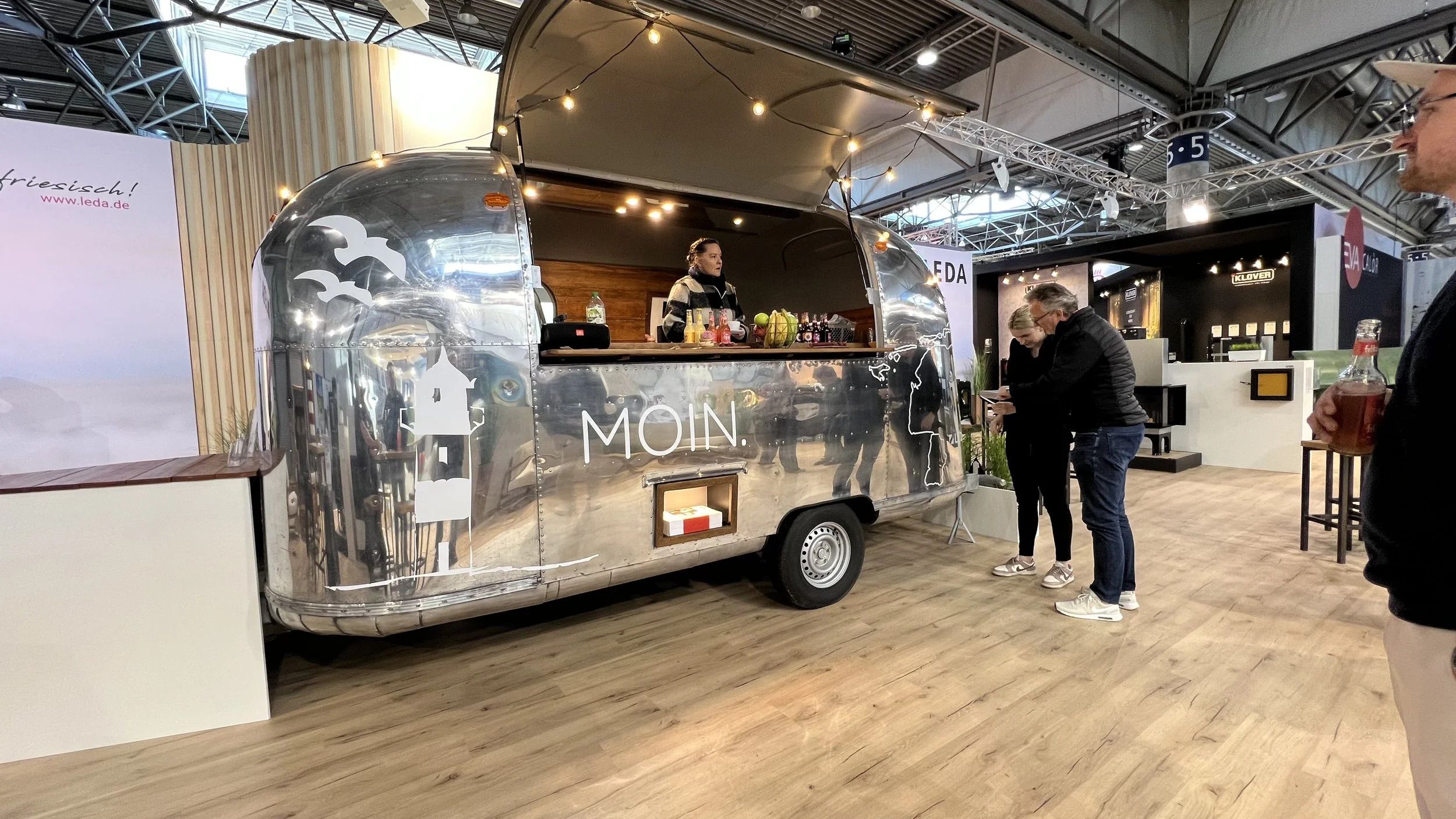 Ein silberner, reflektierender Foodtrailer mit der Aufschrift 'MOIN' am Messestand. Im Inneren verkauft eine Frau Getränke, draußen stehen ein Mann und zwei Frauen beim Kauf und Gespräch. Es ist ein Messegelände mit Holzfußboden und mehreren Ausstellungsständen.