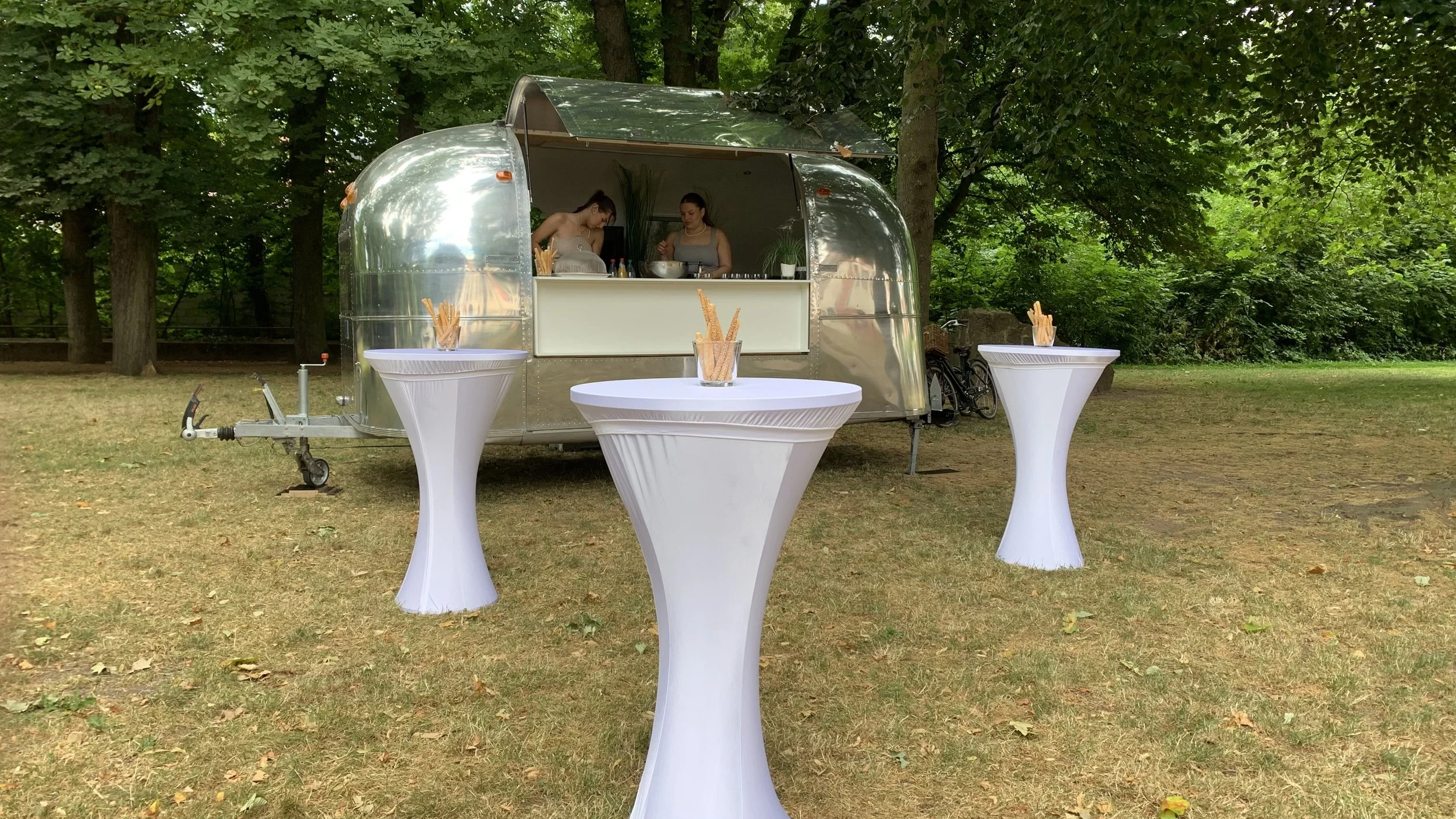 Ein Food-Truck mit einer silbernen, reflektierenden Oberfläche in einem Park, hinter dem zwei Frauen in der Küche stehen. Vor dem Truck sind drei hohe Stehtische mit weißen Überzügen, auf denen Papier- oder Plastikstäbchen in Gläsern stehen.