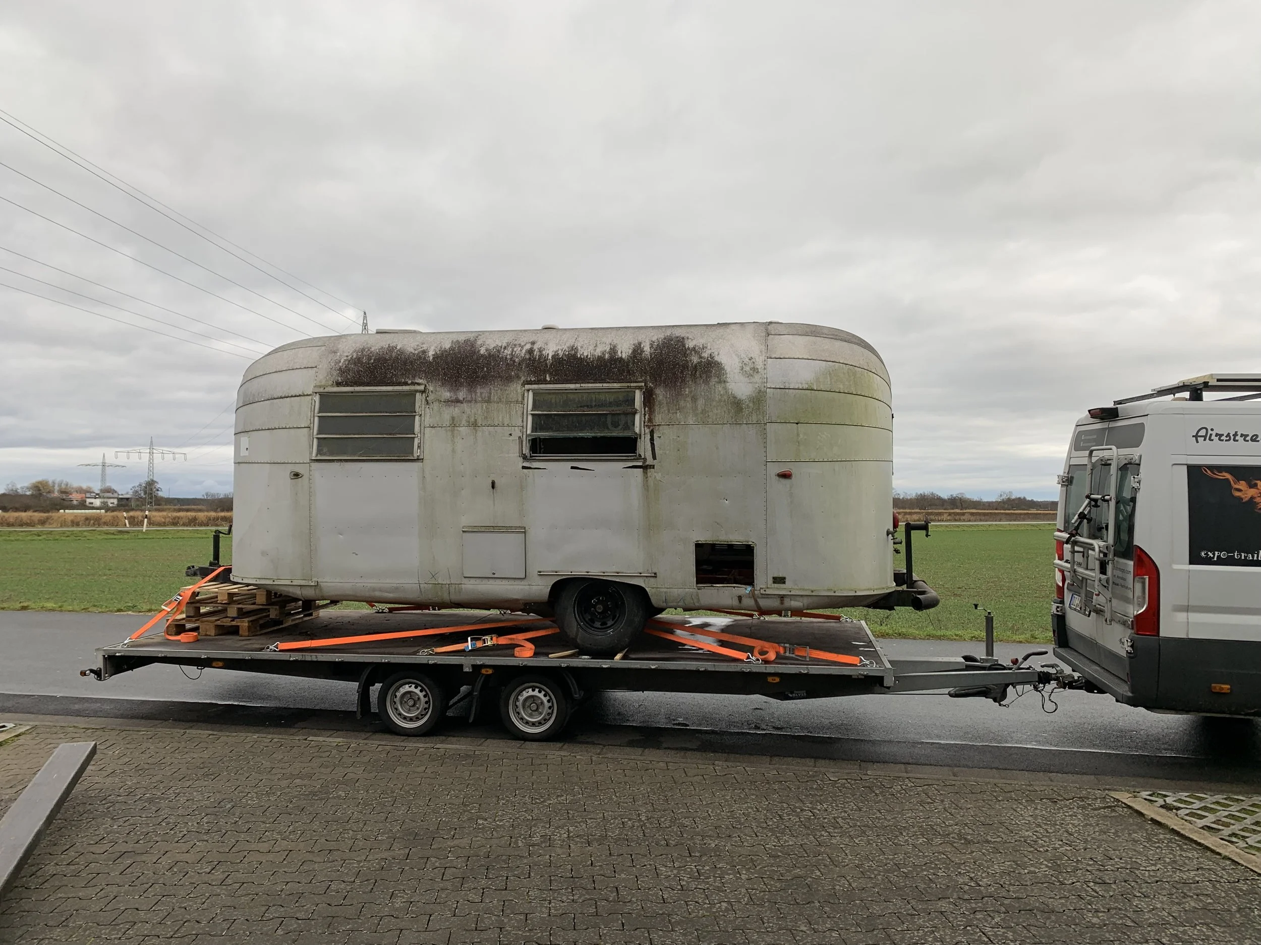 Alte, verlassene Wohnwagen auf einem Transportanhänger mit einem weißen Van im Hintergrund, auf einer Straße in einer ländlichen Gegend bei bewölktem Himmel.
