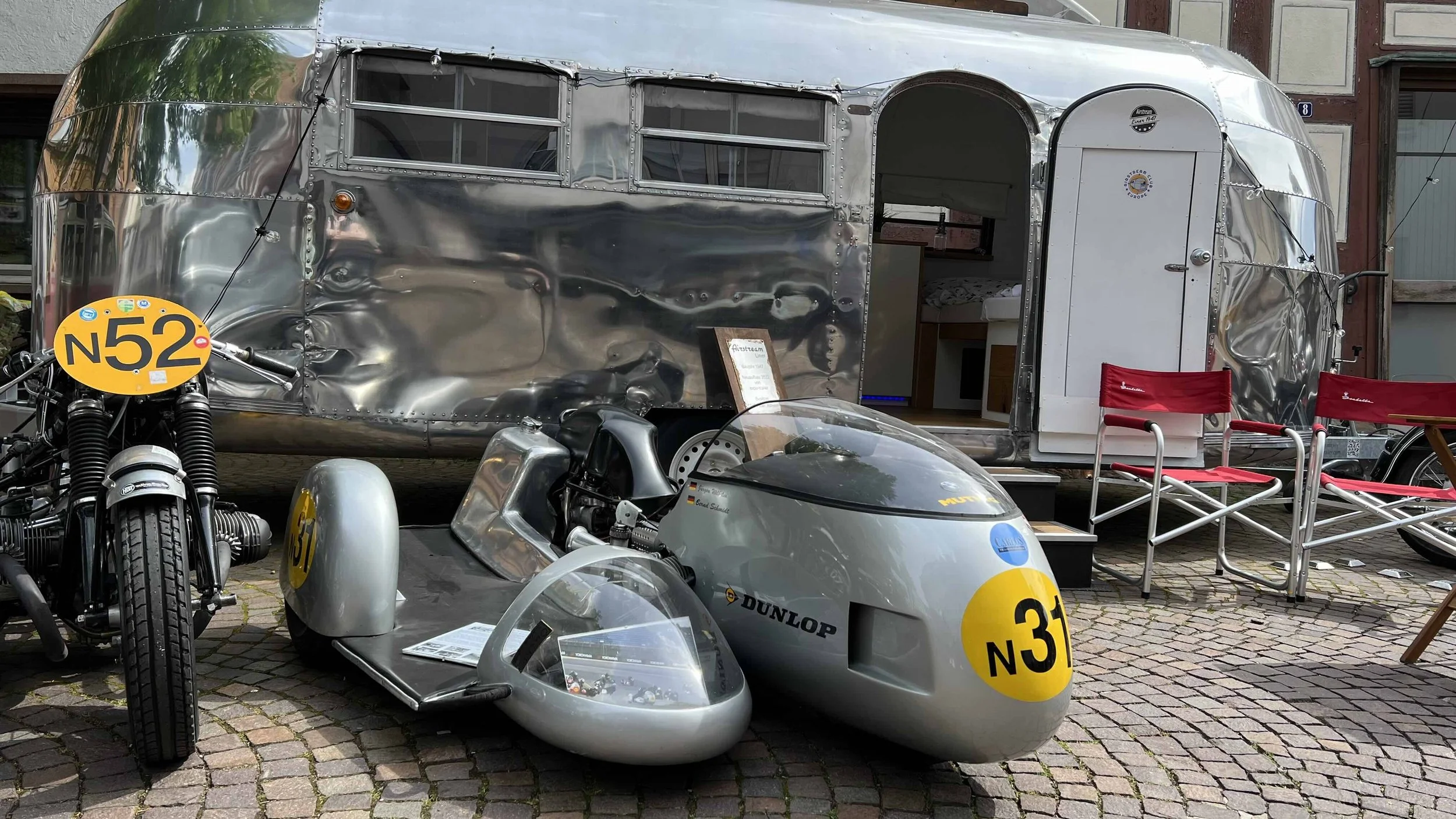 Ein Oldtimer-Rennwagen mit der Nummer 31, ein solothurner Motorrad mit der Nummer N52, eine silberne Airstream-Reisewohnung und einige Erwachsene-Sitze im Freien auf gepflastertem Boden.