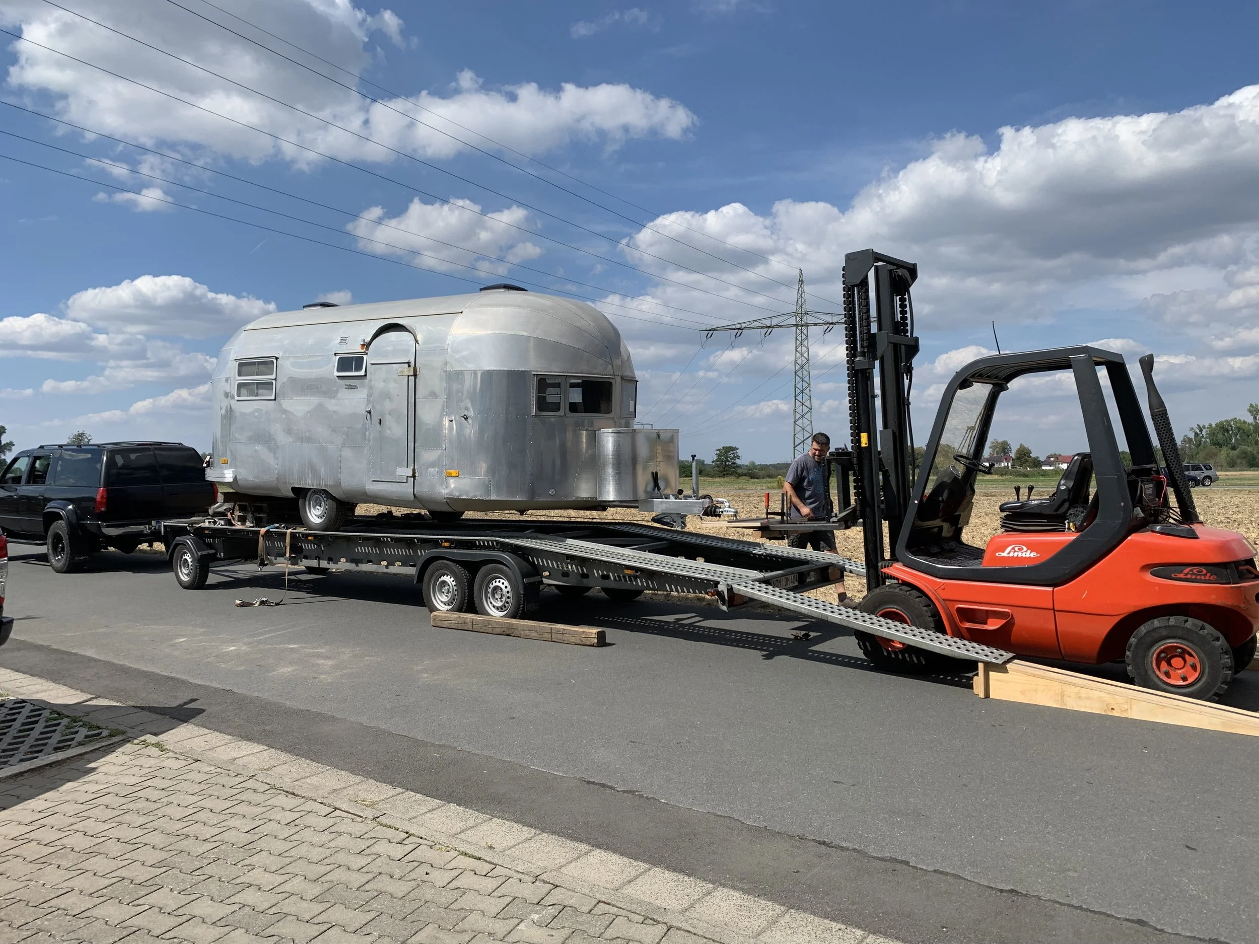Ein silberner Wohnwagen wird mit einem Gabelstapler auf einen Transporter geladen, während eine Person zuschaut.