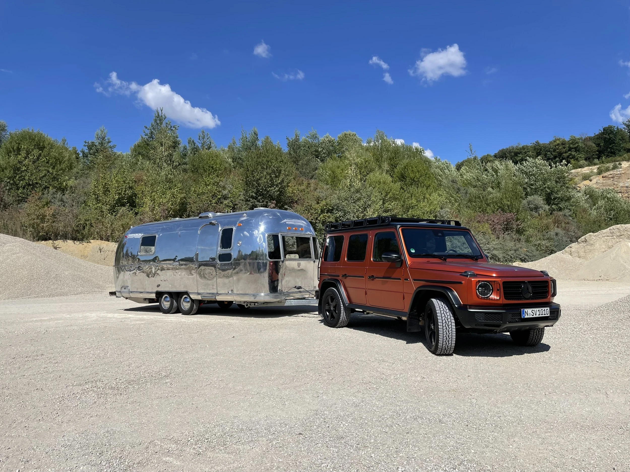 Ein roter Mercedes-Benz G-Class SUV zieht einen silbernen Airstream Wohnwagen auf einem freien, sandigen Gelände mit Baumreihen im Hintergrund und einem blauen Himmel mit Wolken.