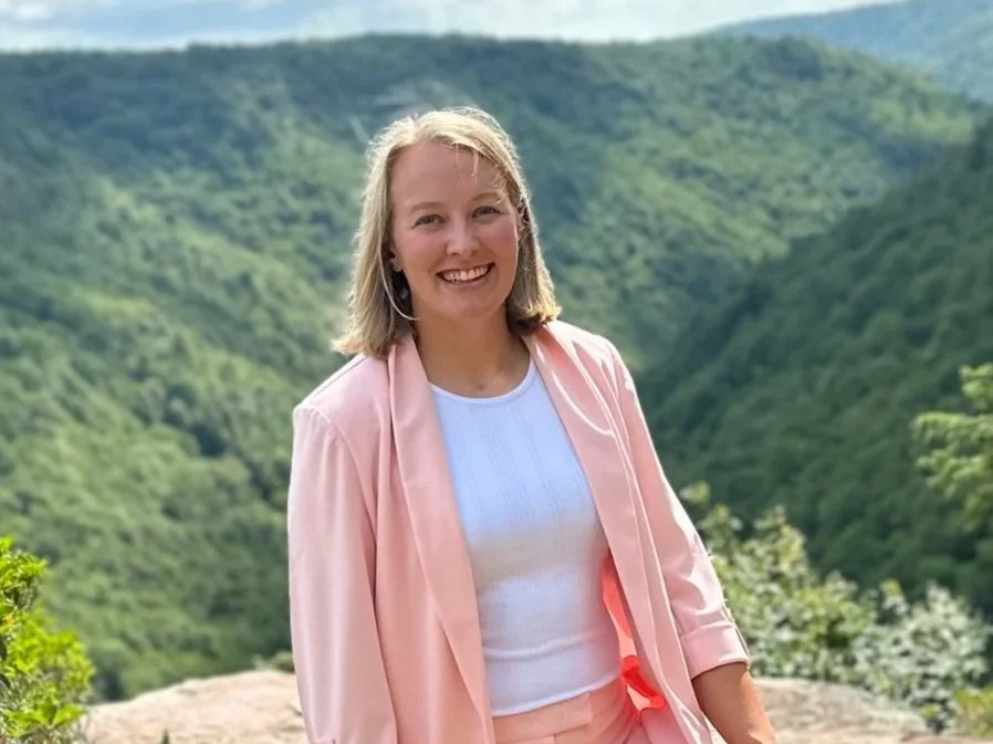 A smiling woman with blonde hair, wearing a pink jacket and white top, standing outdoors with green mountains in the background.