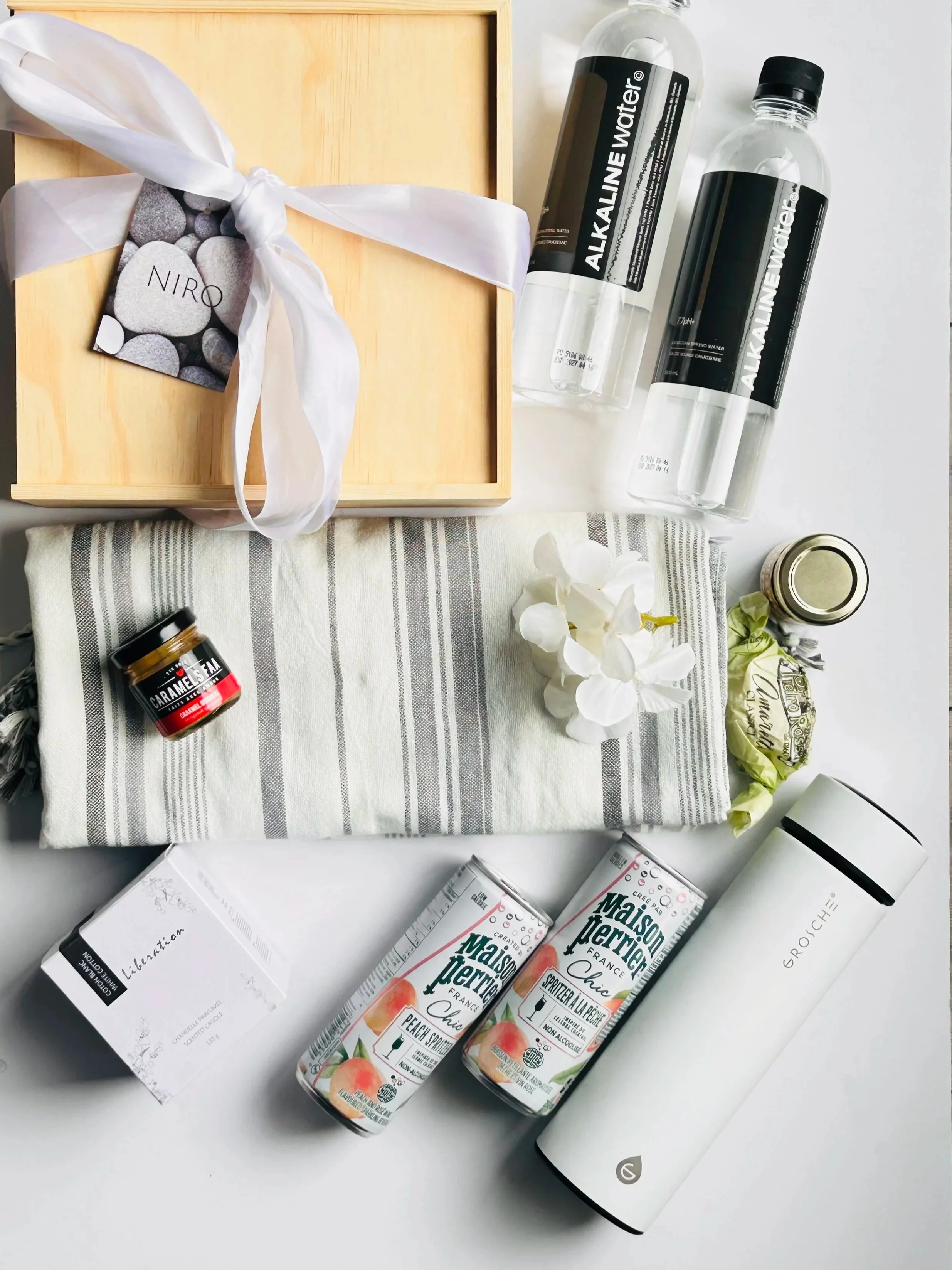 An assortment of items including a wooden box with a white ribbon tied around it, a card with gray rocks and the word 'NIRÖ' on it, two bottles of alkaline water, a jar labeled 'Caramel', a box of peach almond peach spritzer drinks, a white cylindric