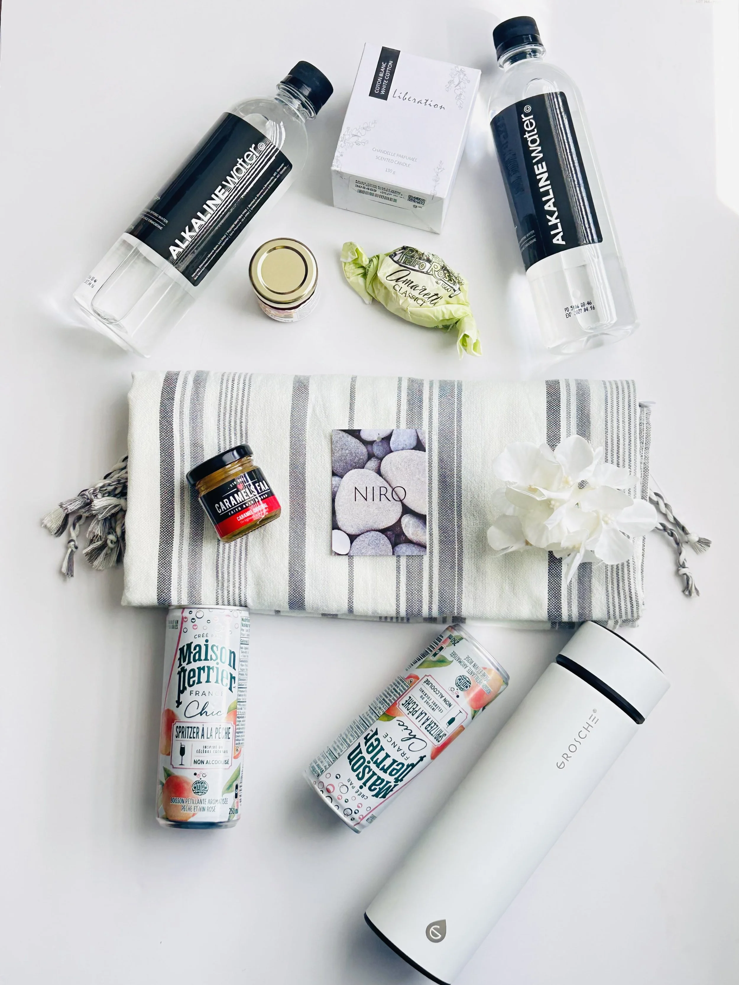 A collection of household items on a white surface, including two bottles of alkaline water, a box of scented candles labeled 'Liberation,' a small jar, a wrapped item, a towel, a white portable speaker, two cans of flavored sparkling water, and a wh