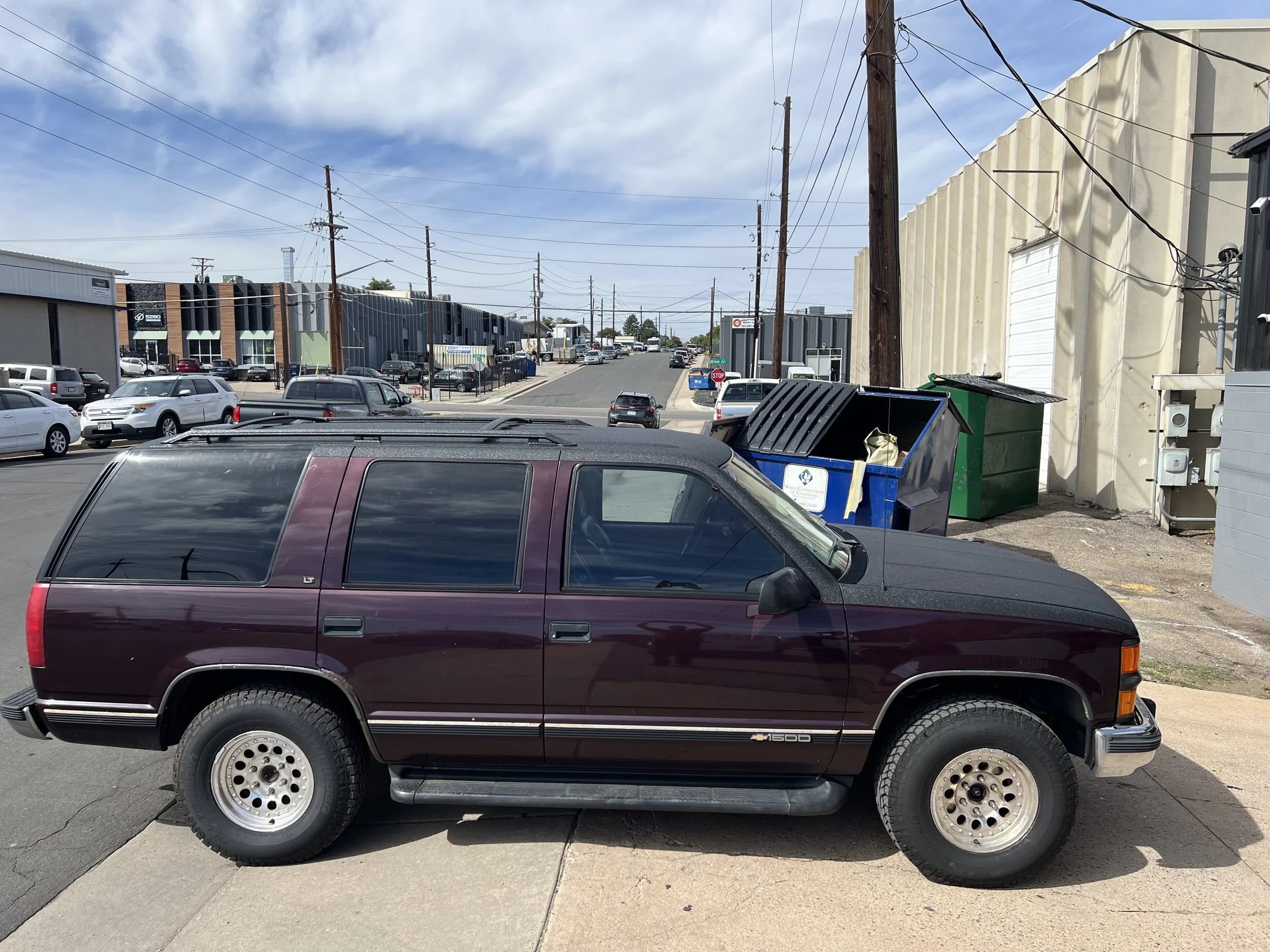 Maroon Jeep Grand Cherokee parked on sidewalk on a city street with commercial buildings in the background and garbage dumpsters behind it.