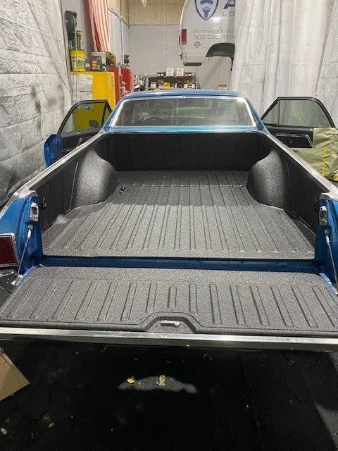 The image shows the bed of a pickup truck with Mil+Spec Liner sprayed on the bed and the tailgate inside workshop. The truck's cabin is visible in the background, with two open doors on either side.
