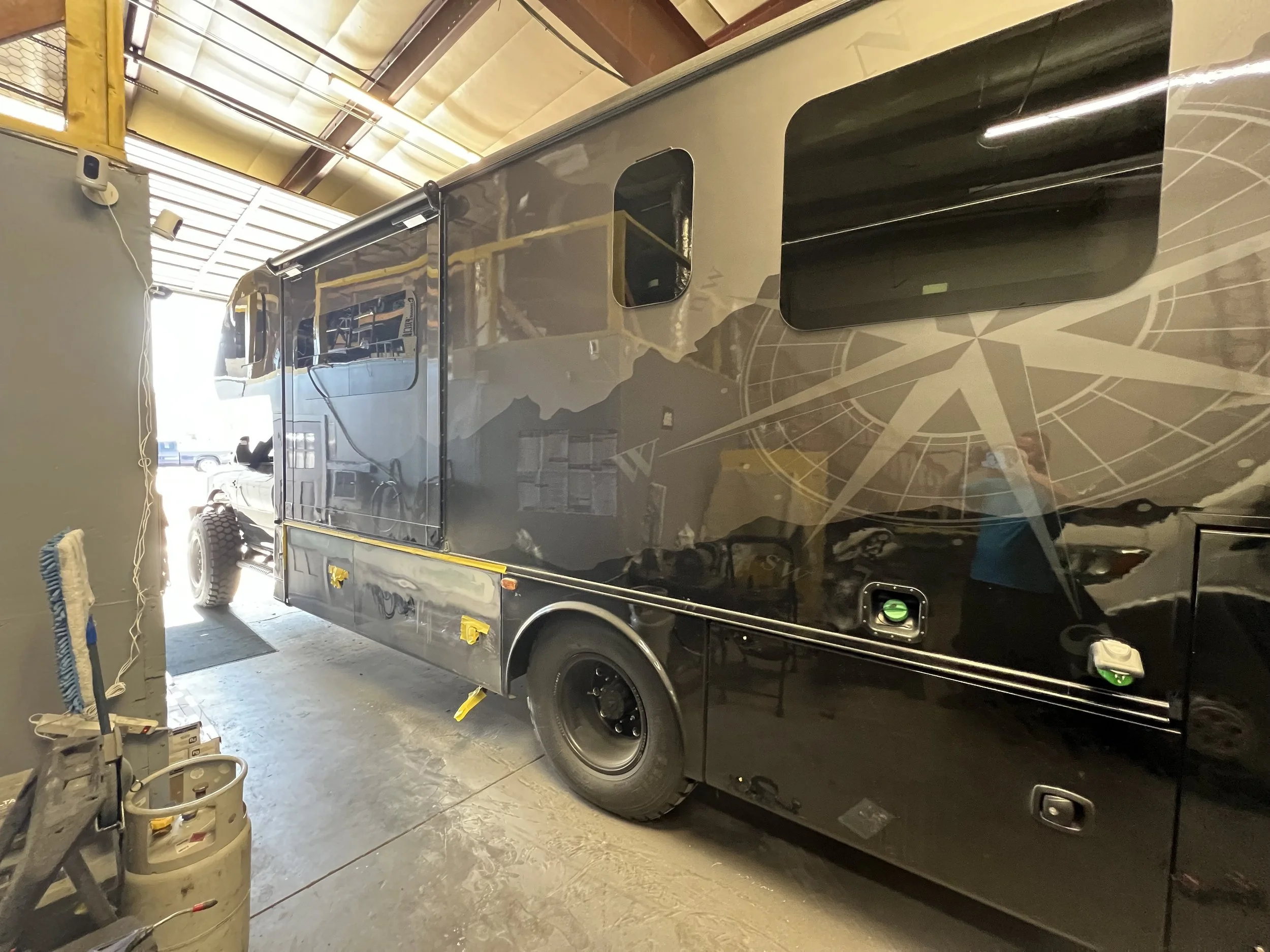 Black RV parked inside a warehouse with tools and supplies nearby.
