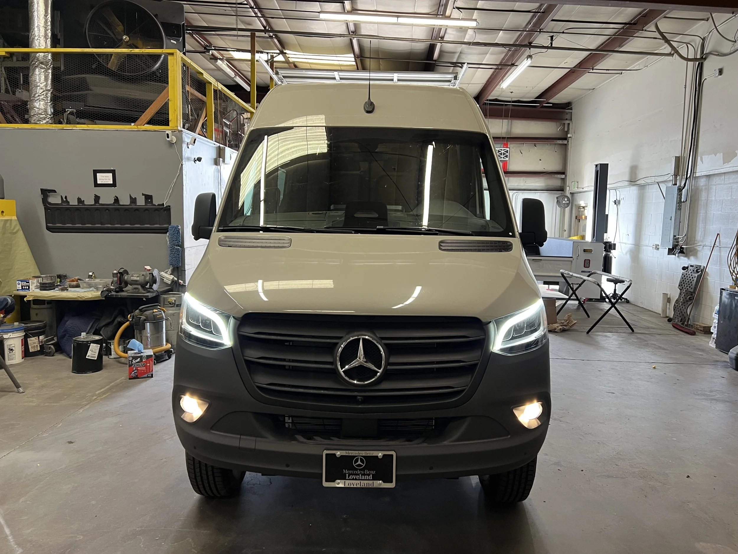 A white Mercedes-Benz van inside a garage, front view with headlights on, black grille, Mercedes logo, license plate, and various tools and equipment in the background.