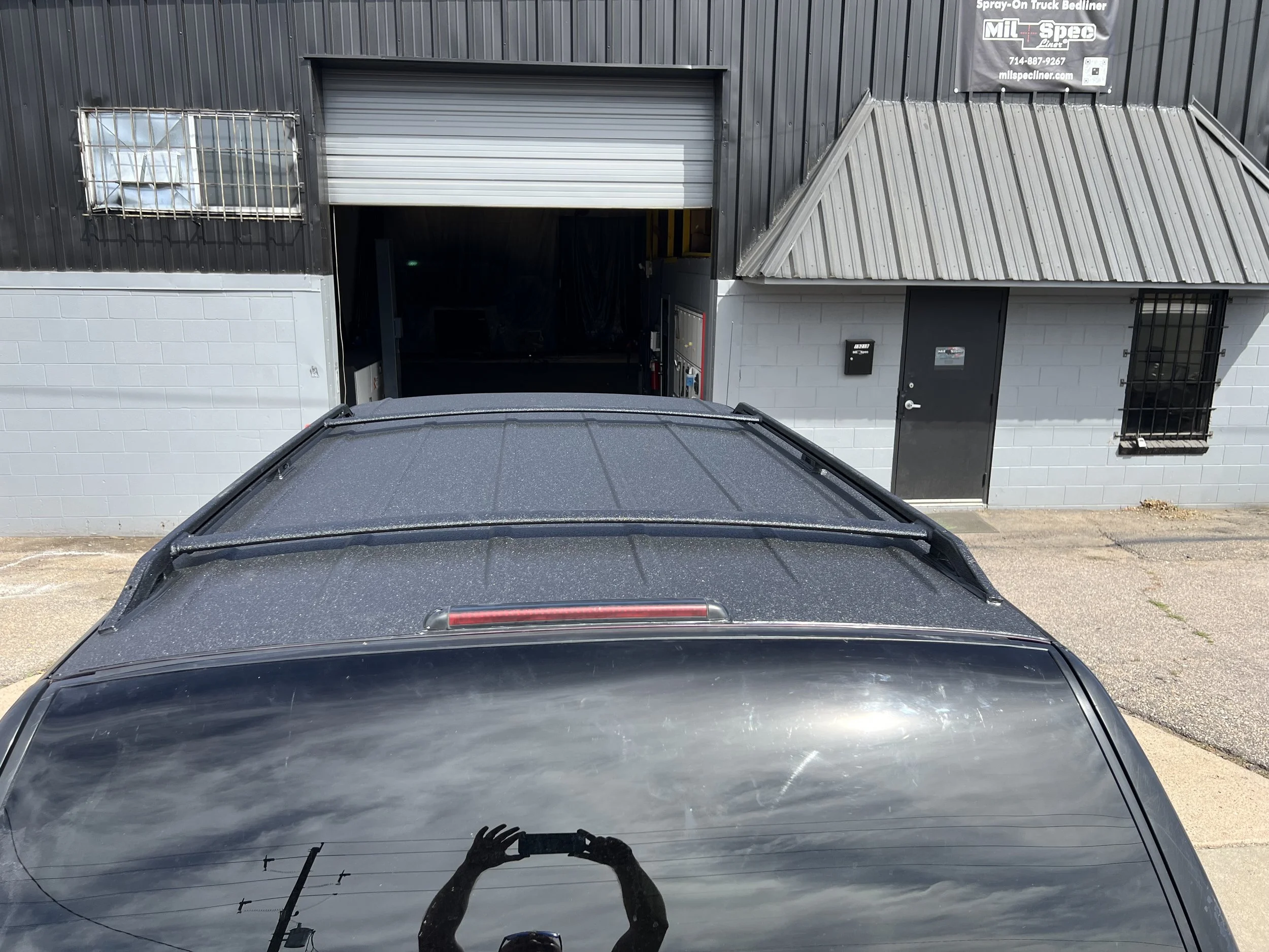 Black vehicle with a dusty roof parked outside a building with a gray and black exterior. The building has a small window with security bars, a black door, and an open garage door revealing the interior. There is a person taking a photo reflected in 