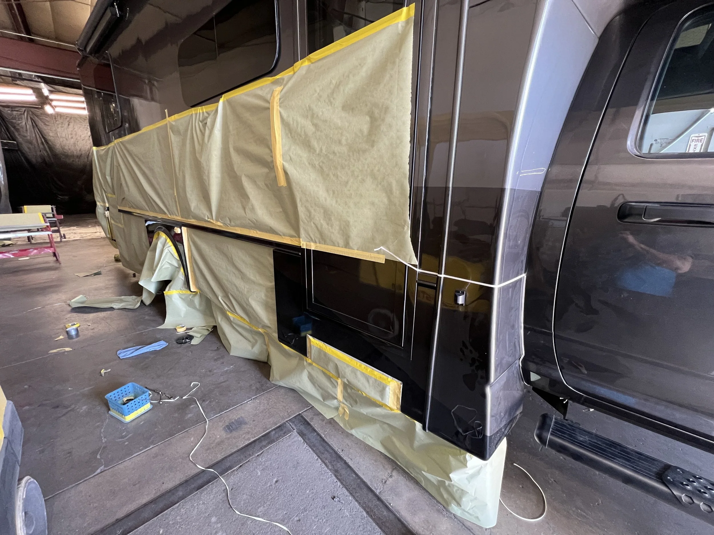A black vehicle is covered with protective paper and tape, likely for painting or repair work, inside a workshop or garage.