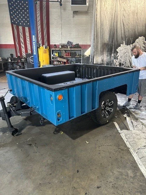 A small, blue, custom-built trailer with a black interior, parked inside a workshop. A man is working beside it, and tools are visible on shelves in the background.