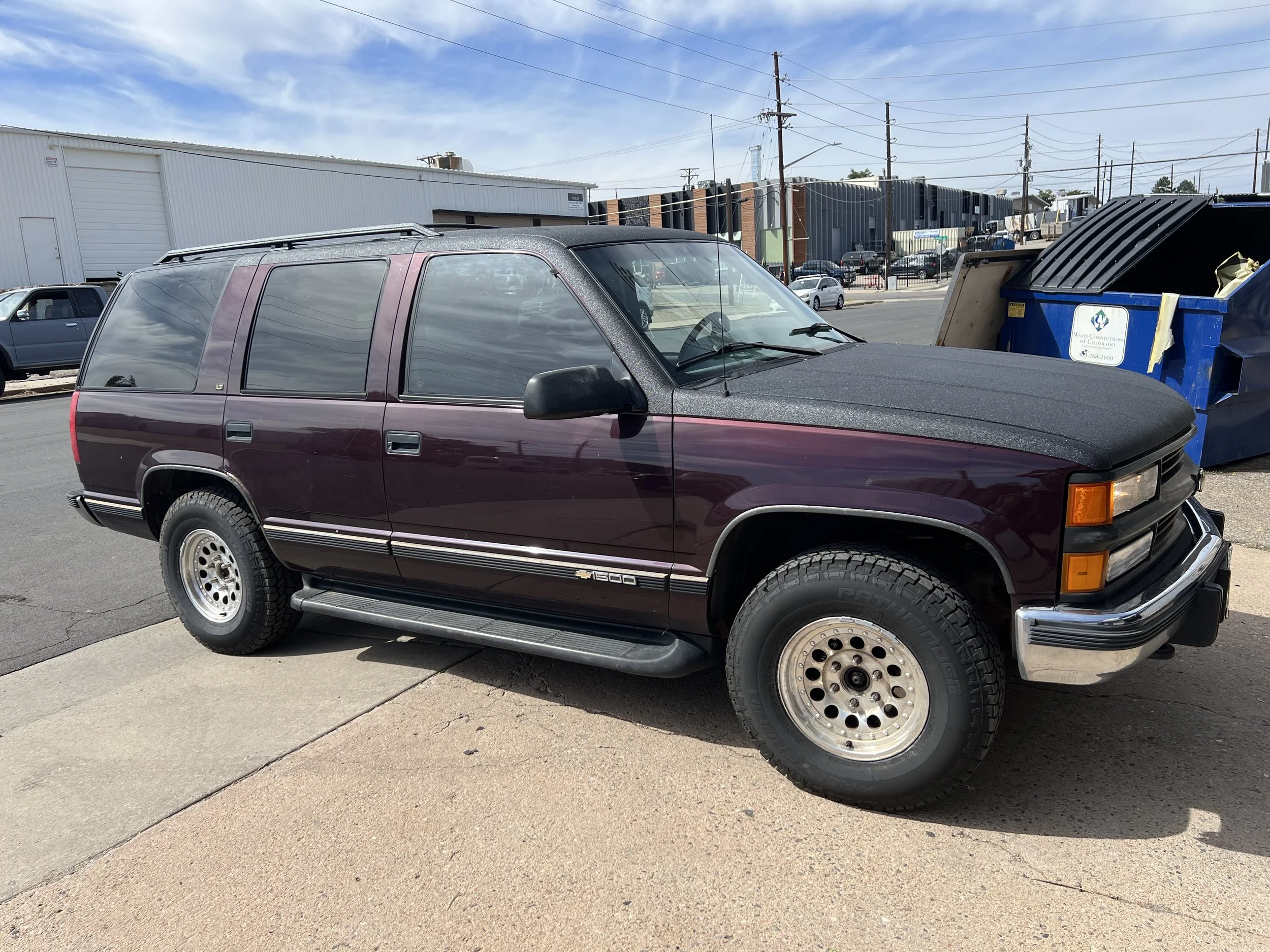 A vintage mid-1990s Chevrolet SUV parked on a paved street with a large industrial building and a blue dumpster in the background.
