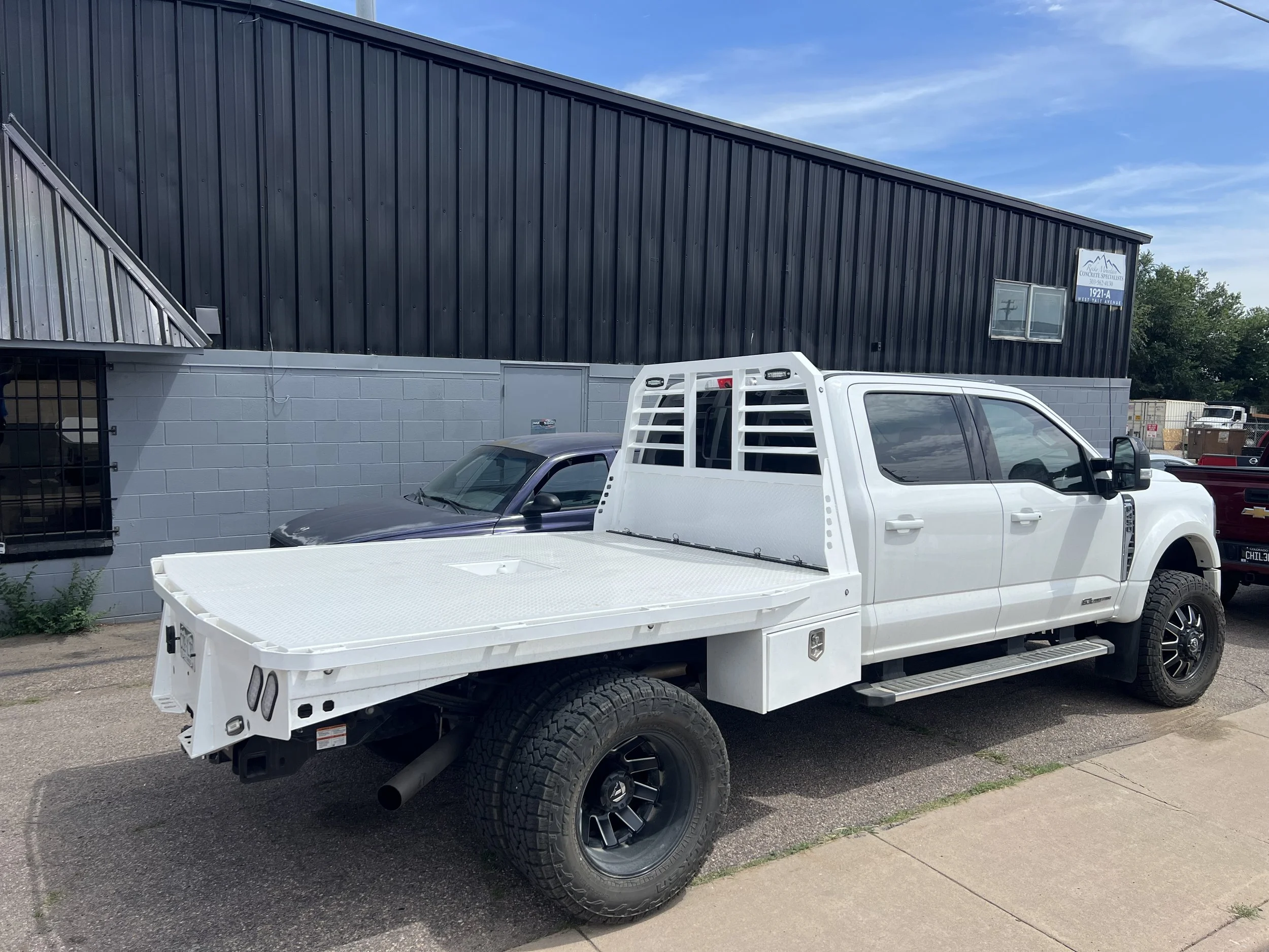 White utility truck with a flatbed parked on a street, with a black building and another vehicle in the background.