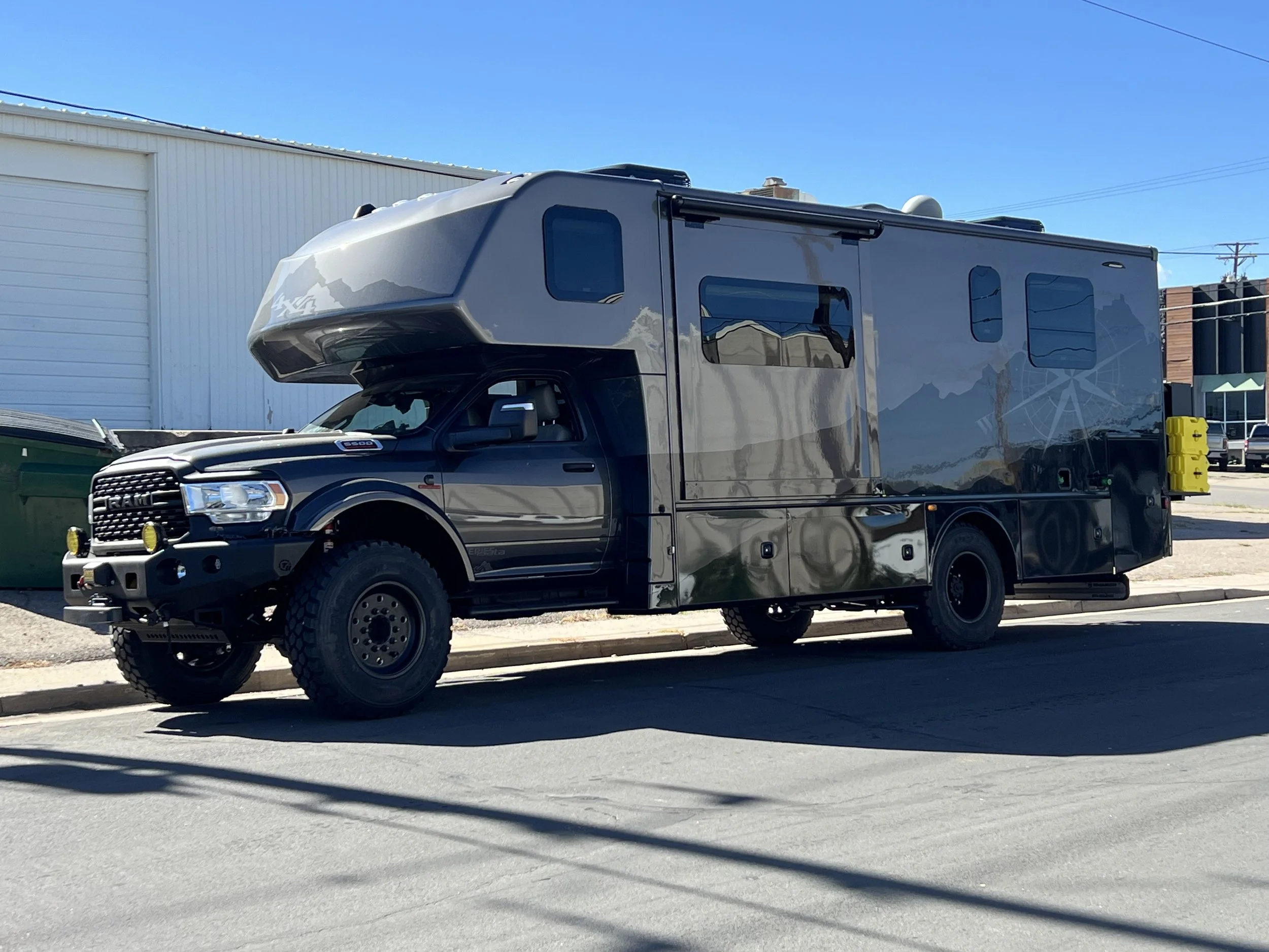 A black camper truck parked on the street under a clear blue sky with a white building in the background.