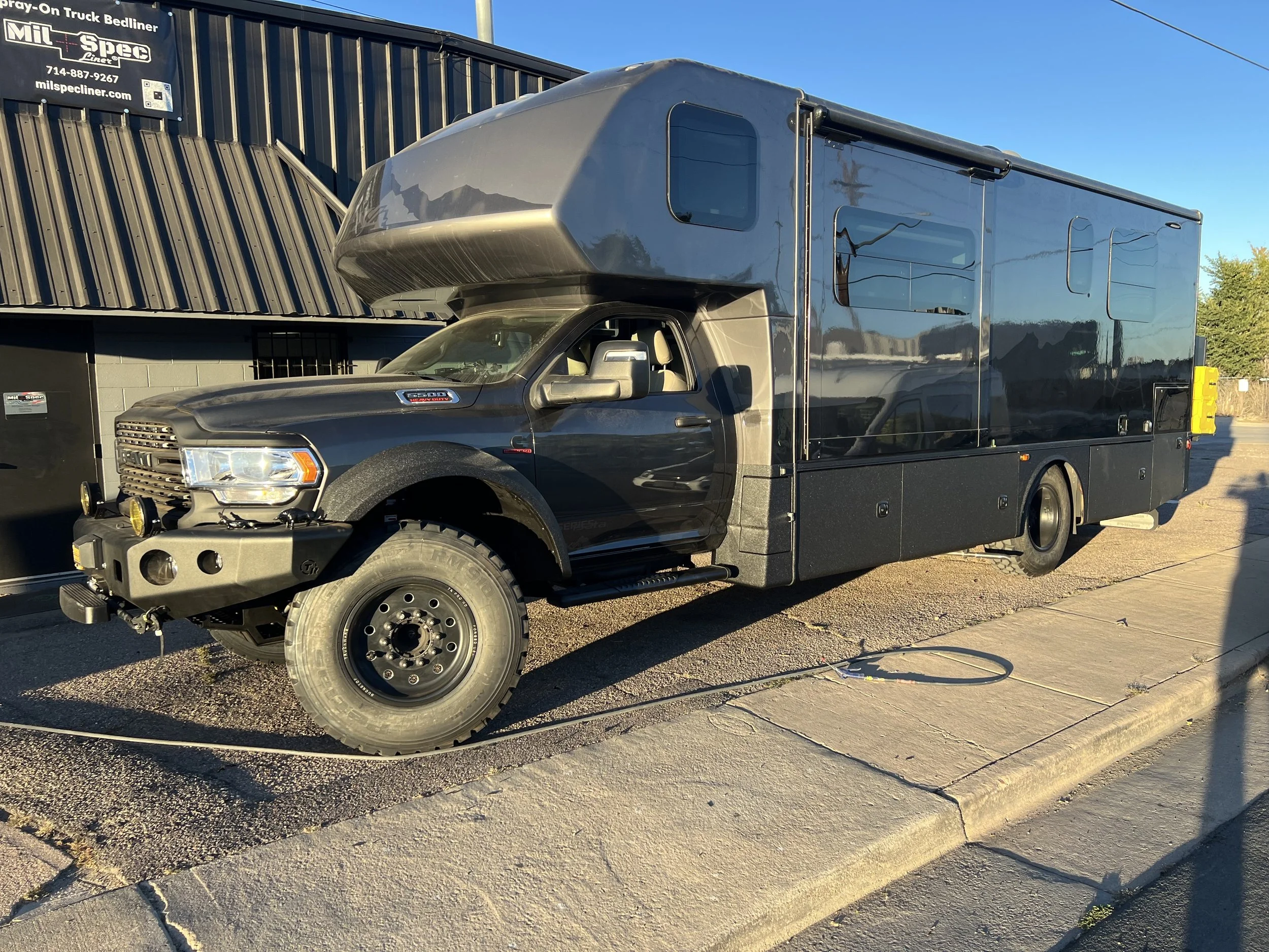 A black emergency response vehicle with a large boxy trailer attached, parked on a sidewalk during sunset.