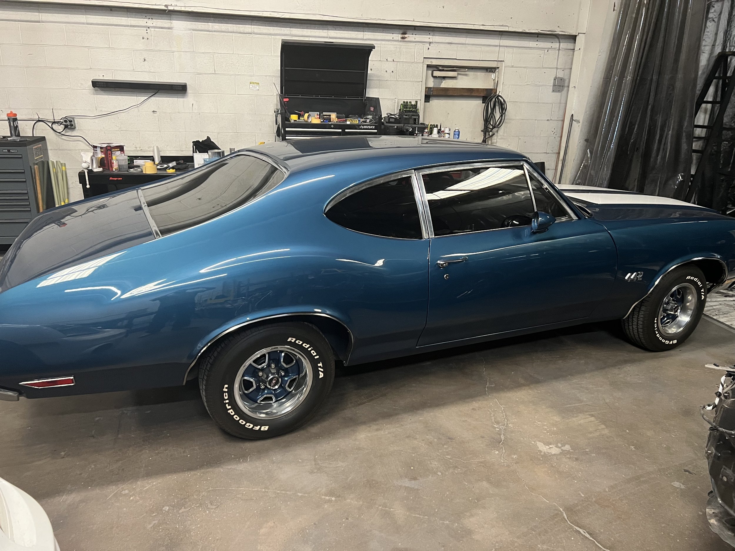 A vintage blue coupe car with a rear glass bubble window, parked inside a garage with tools and equipment on shelves and a cabinet in the background.