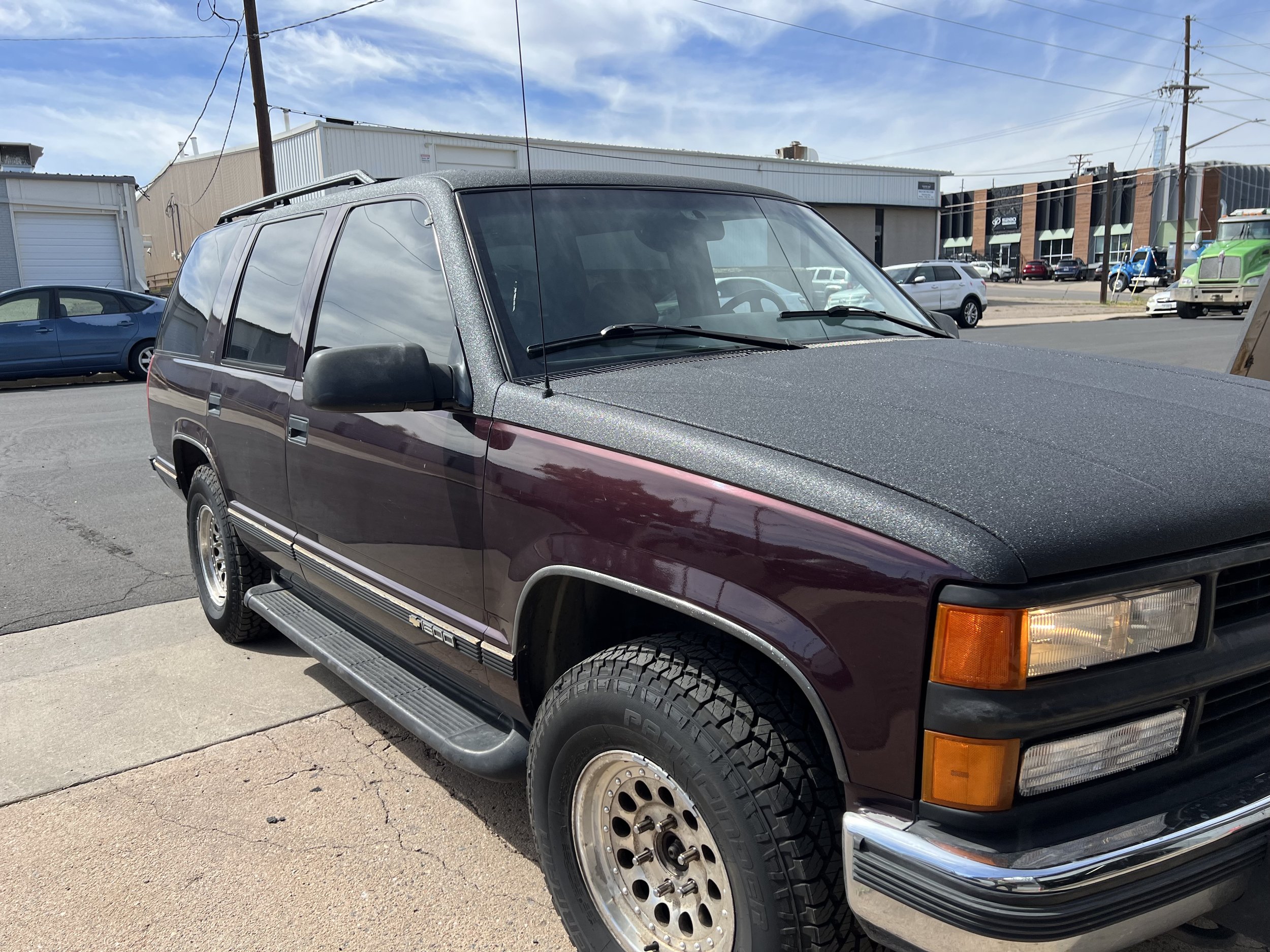 A maroon Chevrolet SUV with black accents parked on a sidewalk during daytime, with street and commercial buildings in the background.