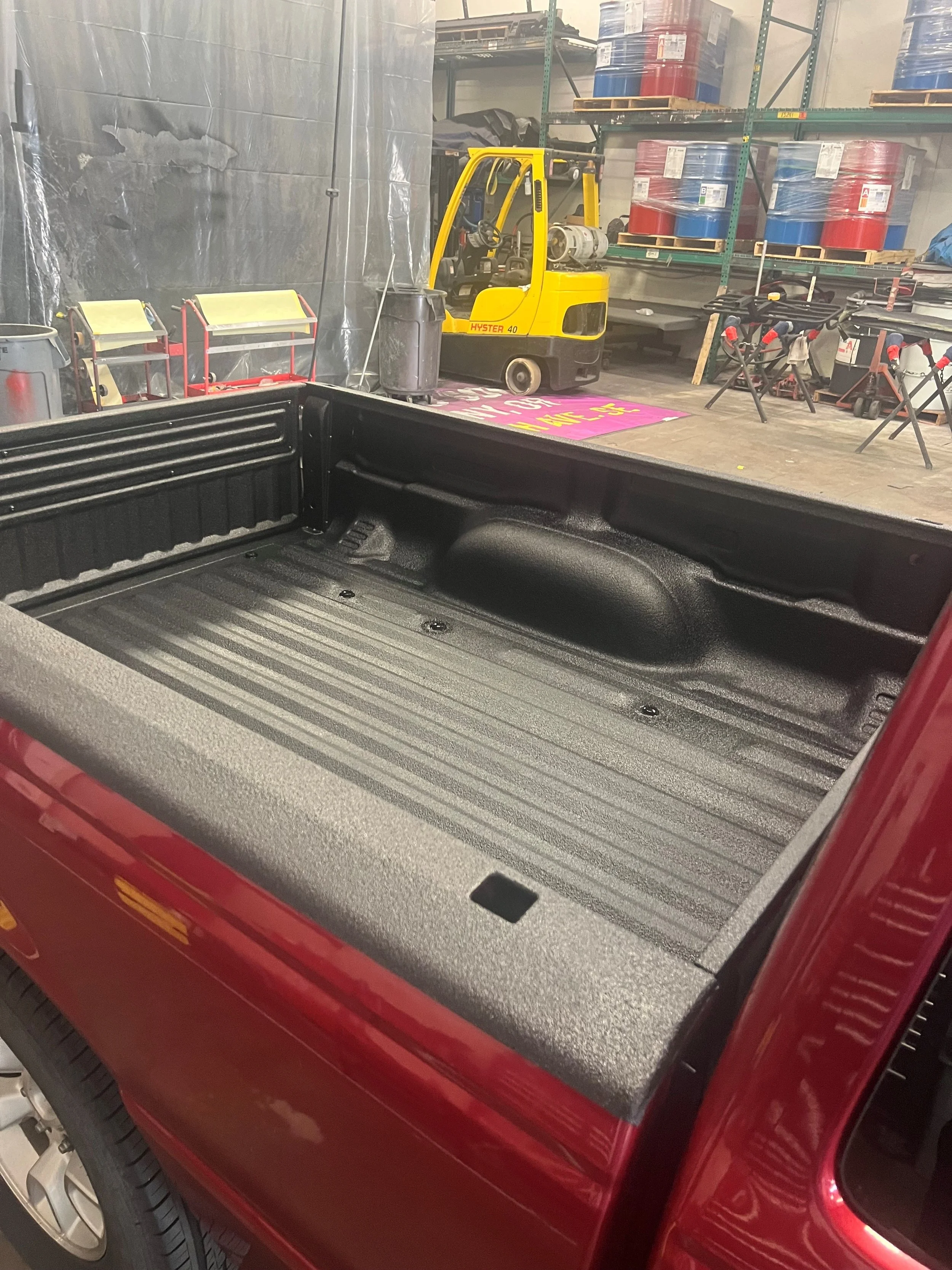 The image shows a red pickup truck bed with a textured black Mil+Spec Spray-on Bedliner, inside a workshop.