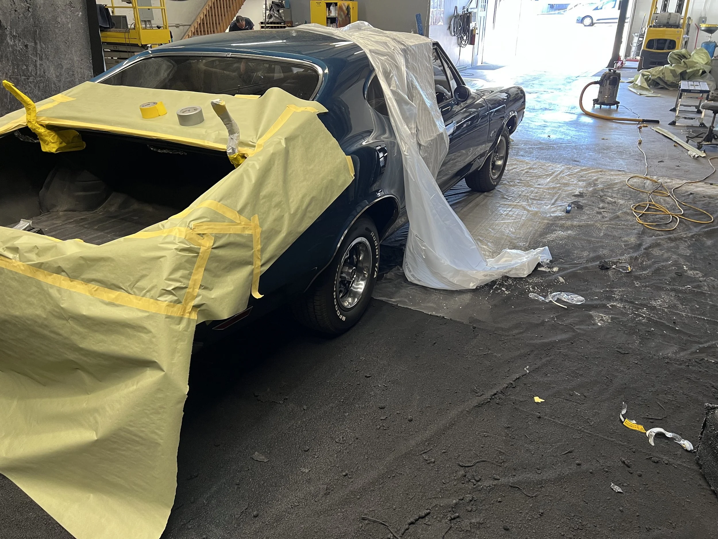 A classic blue car is being painted or repaired inside a garage, with masking paper and painter's tape applied to various parts of the car, and work tools and equipment visible around.