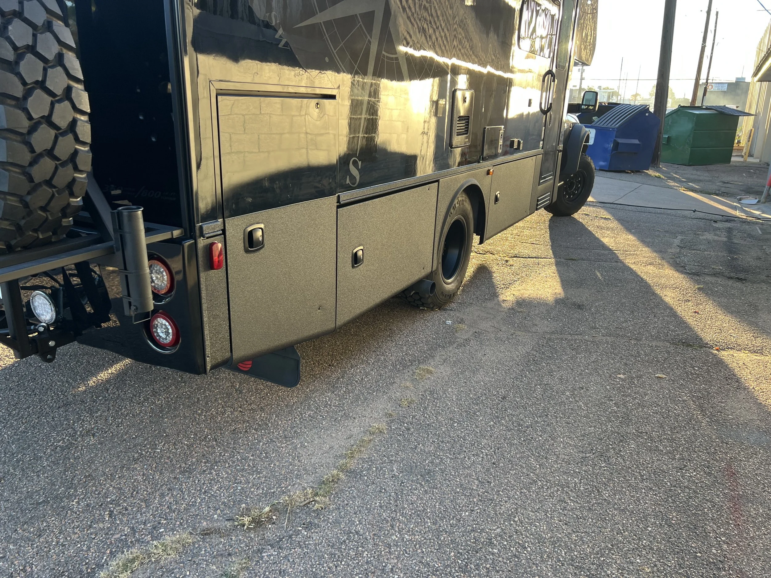 Black utility truck with storage compartments parked on the street during sunset, with green and blue trash bins in the background.
