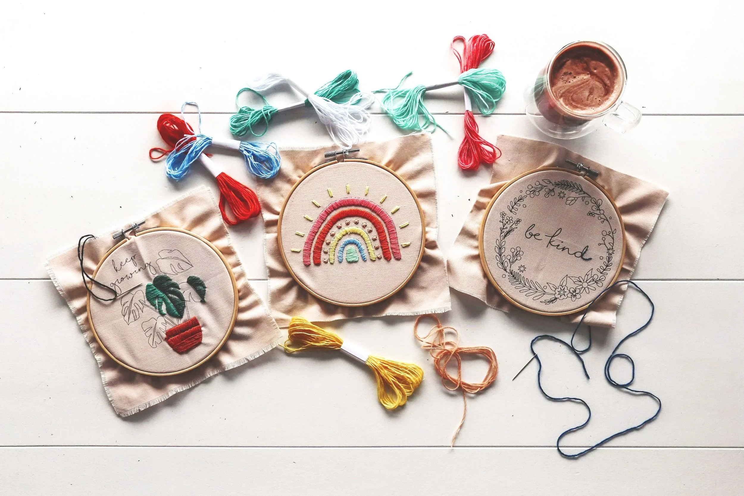 Embroidery hoops with colorful designs and messages, embroidery floss, and a mug of hot chocolate on a white wooden surface.