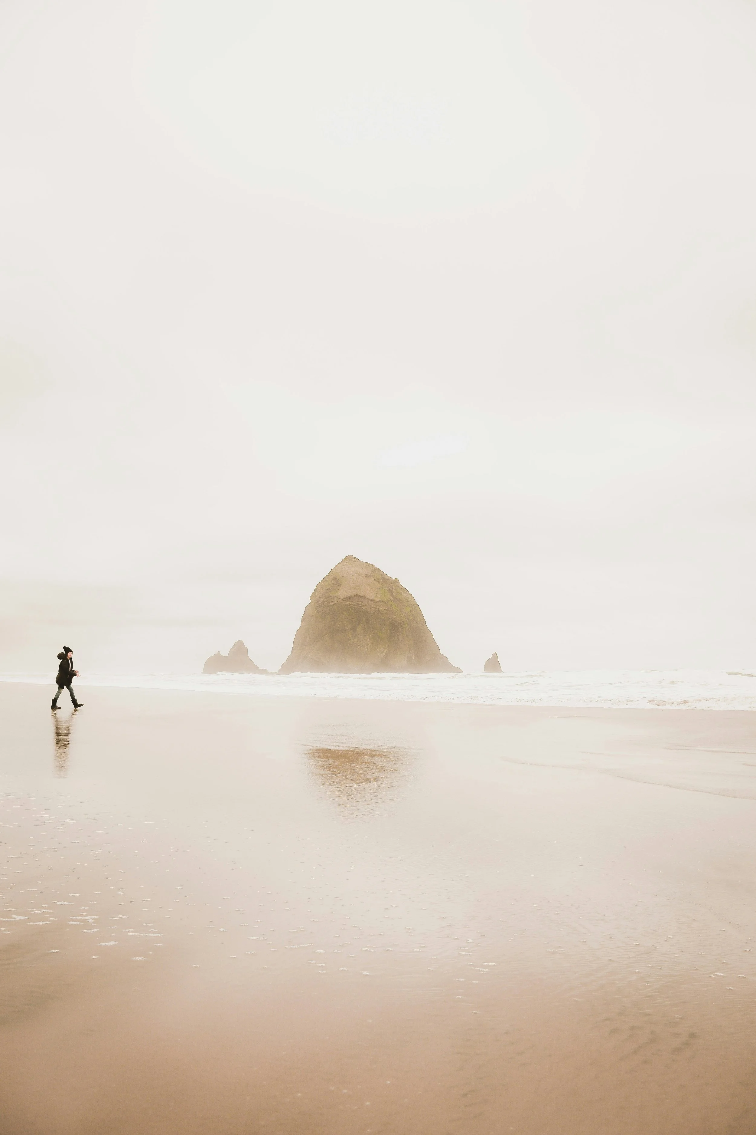 A solitary person walking along a foggy beach near large rock formations in the water.
