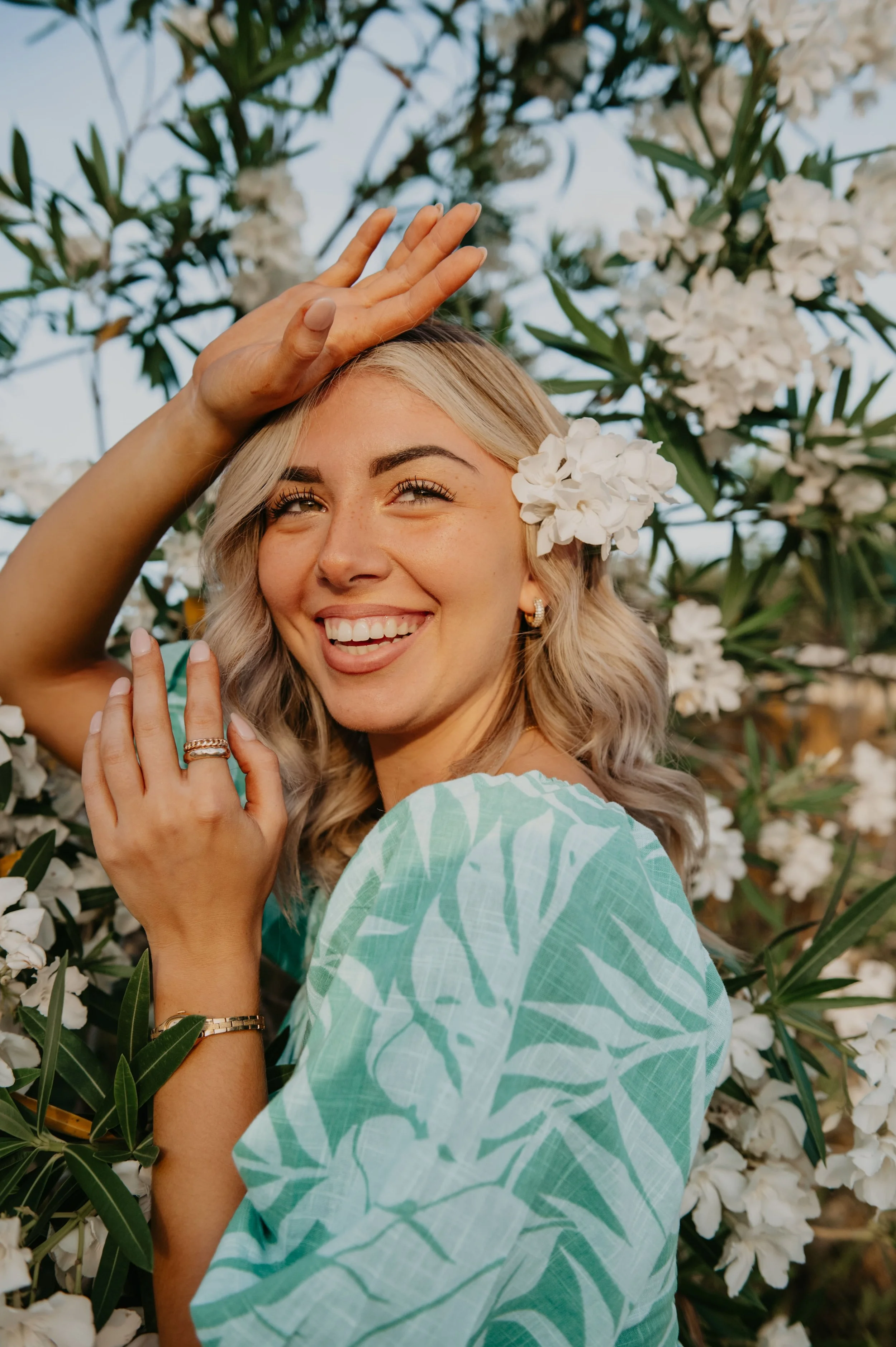 A smiling woman with blonde hair and flowers in her hair, wearing a light green patterned top, standing among white flowering plants.