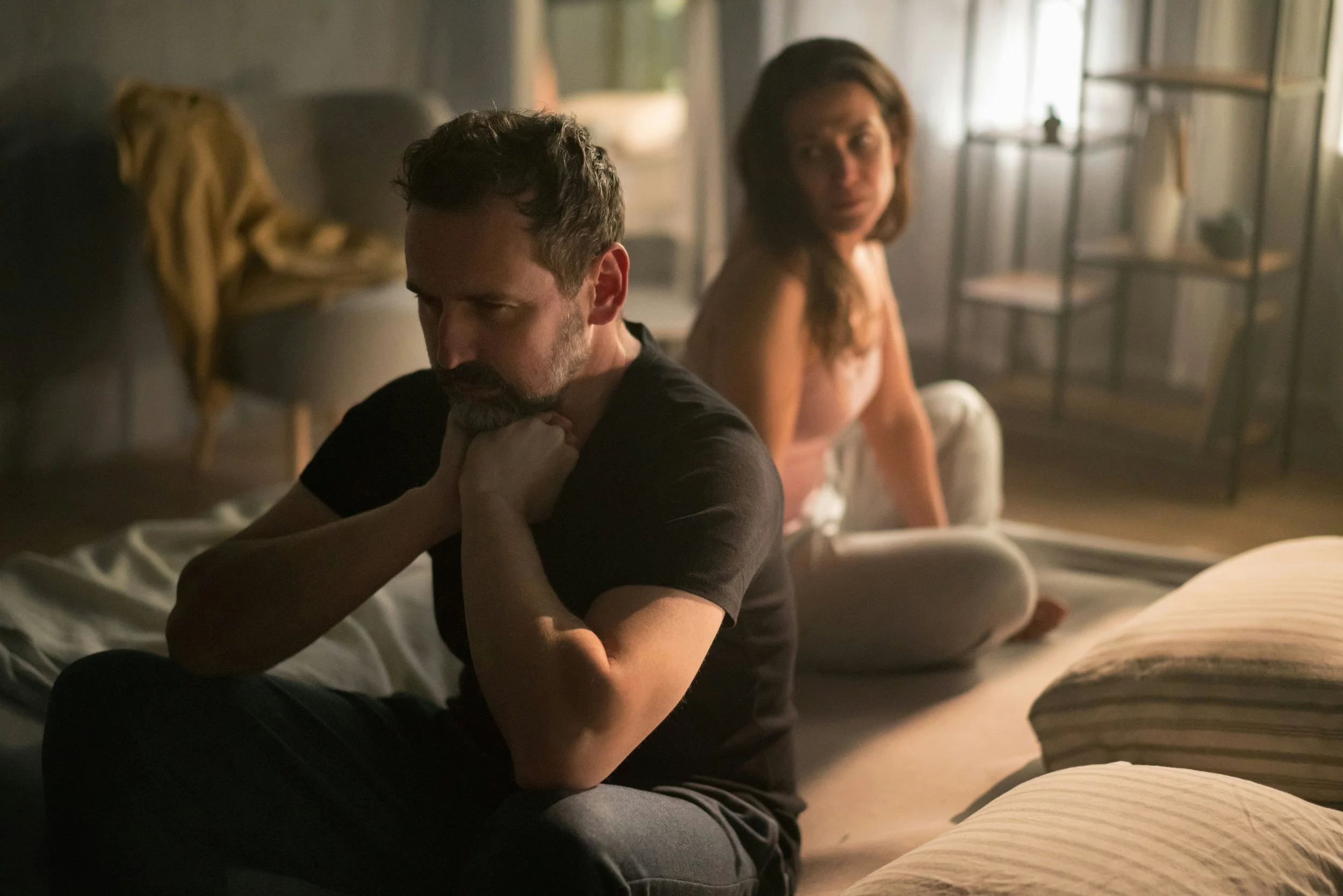 A man and a woman sit on a bed in a dimly lit room. The man appears upset or contemplative, with his hand on his neck, while the woman looks at him with concern or curiosity.