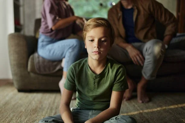 A young boy sitting on the floor with a serious expression, while two adults sit on a couch behind him.