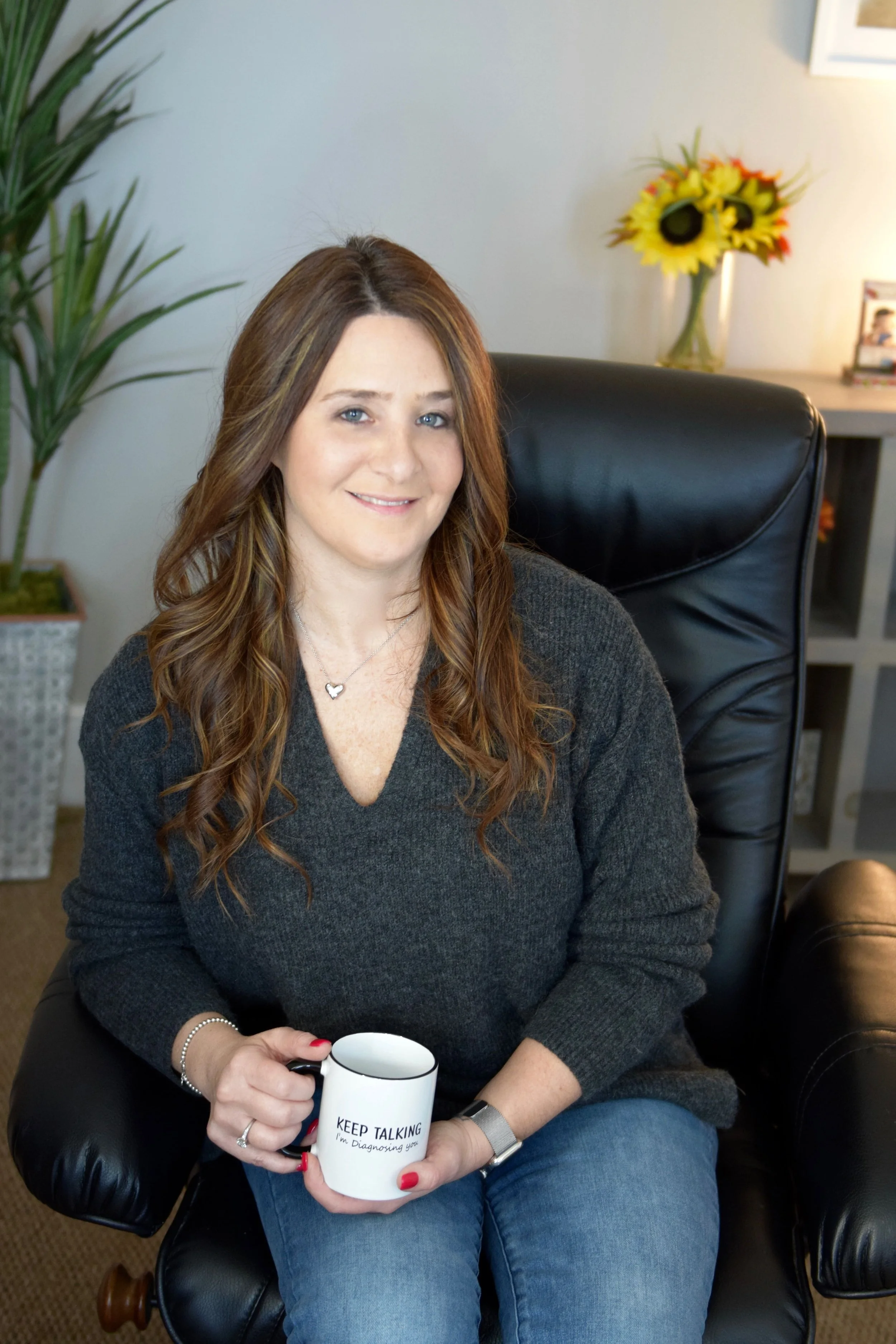 A woman with long, wavy brown hair sitting in a black leather chair, holding a white mug that says 'Keep Talking, I'm Diagnosing You' in an indoor setting with houseplants and sunflower decor.