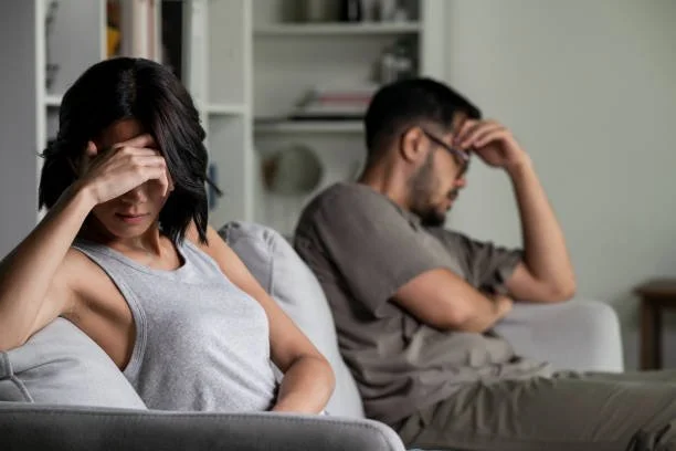 A woman and man sitting on a couch, both appearing upset with their foreheads resting on their hands.