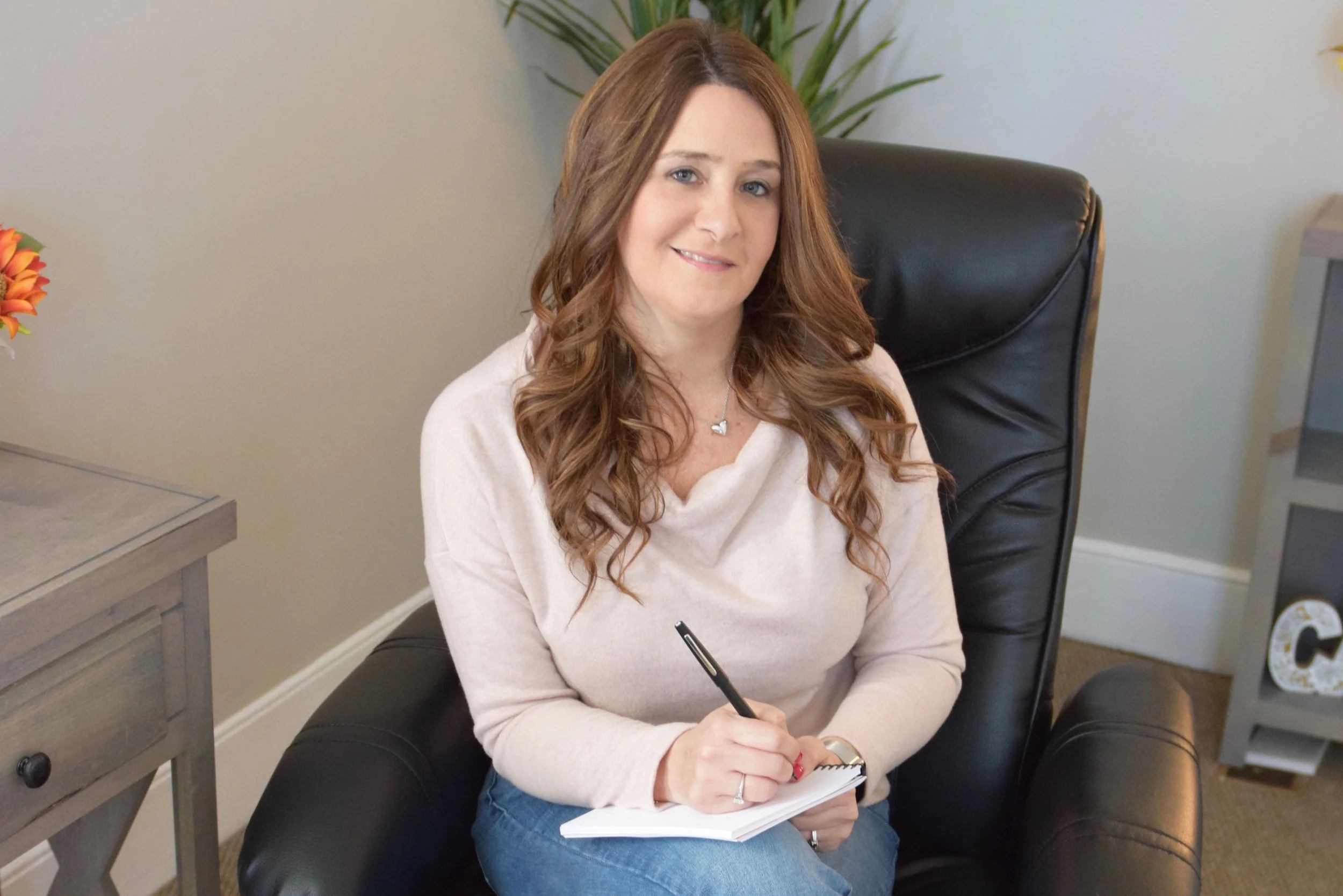 A woman with long, wavy brown hair, light skin, sitting in a black leather office chair, holding a pen and a notepad, smiling at the camera in a well-lit room with a plant in the background.