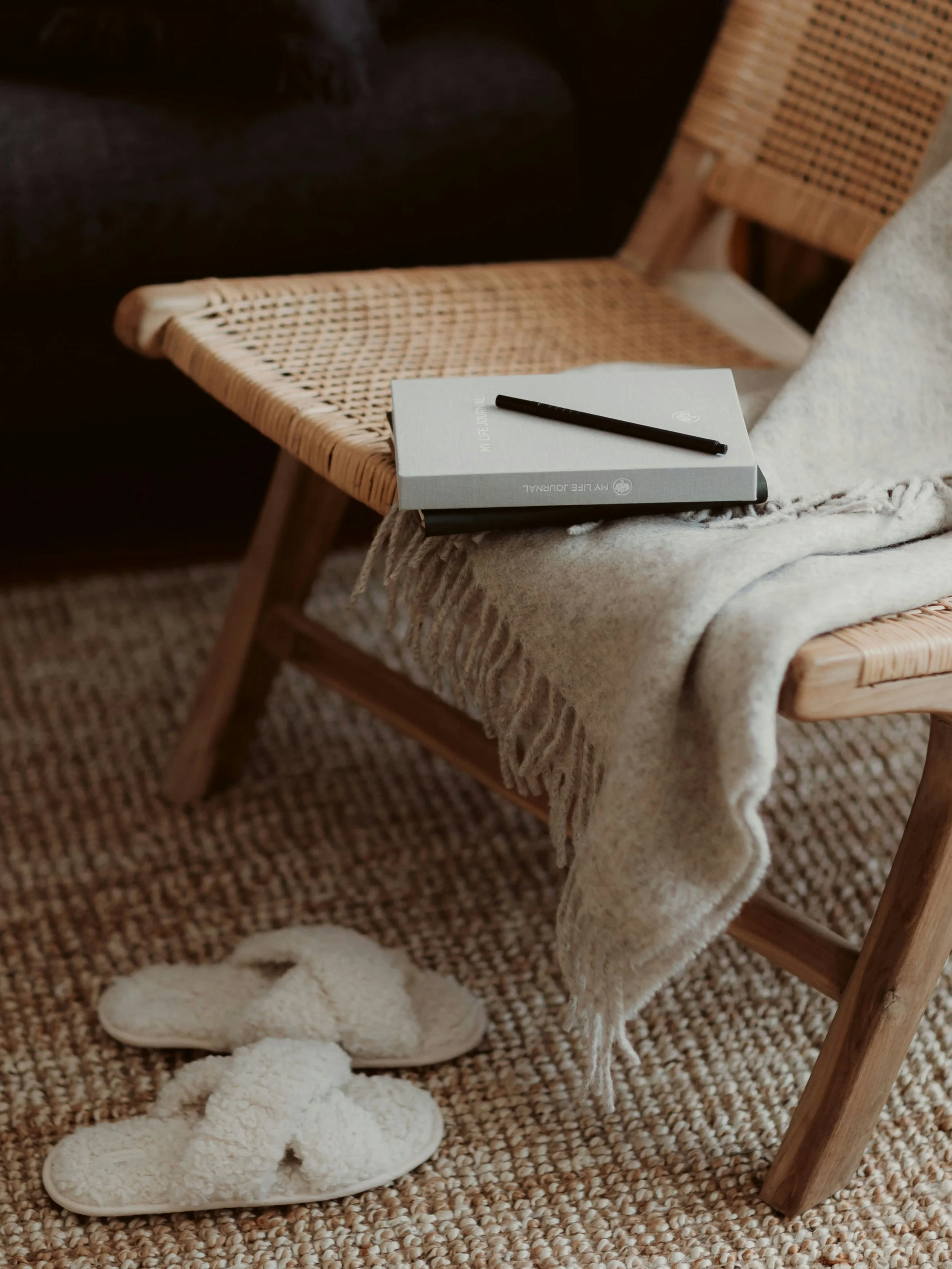 A cozy scene with a woven wicker chair, a beige throw blanket, a notebook with a black pen on top, and a pair of fluffy white slippers on a textured beige rug.