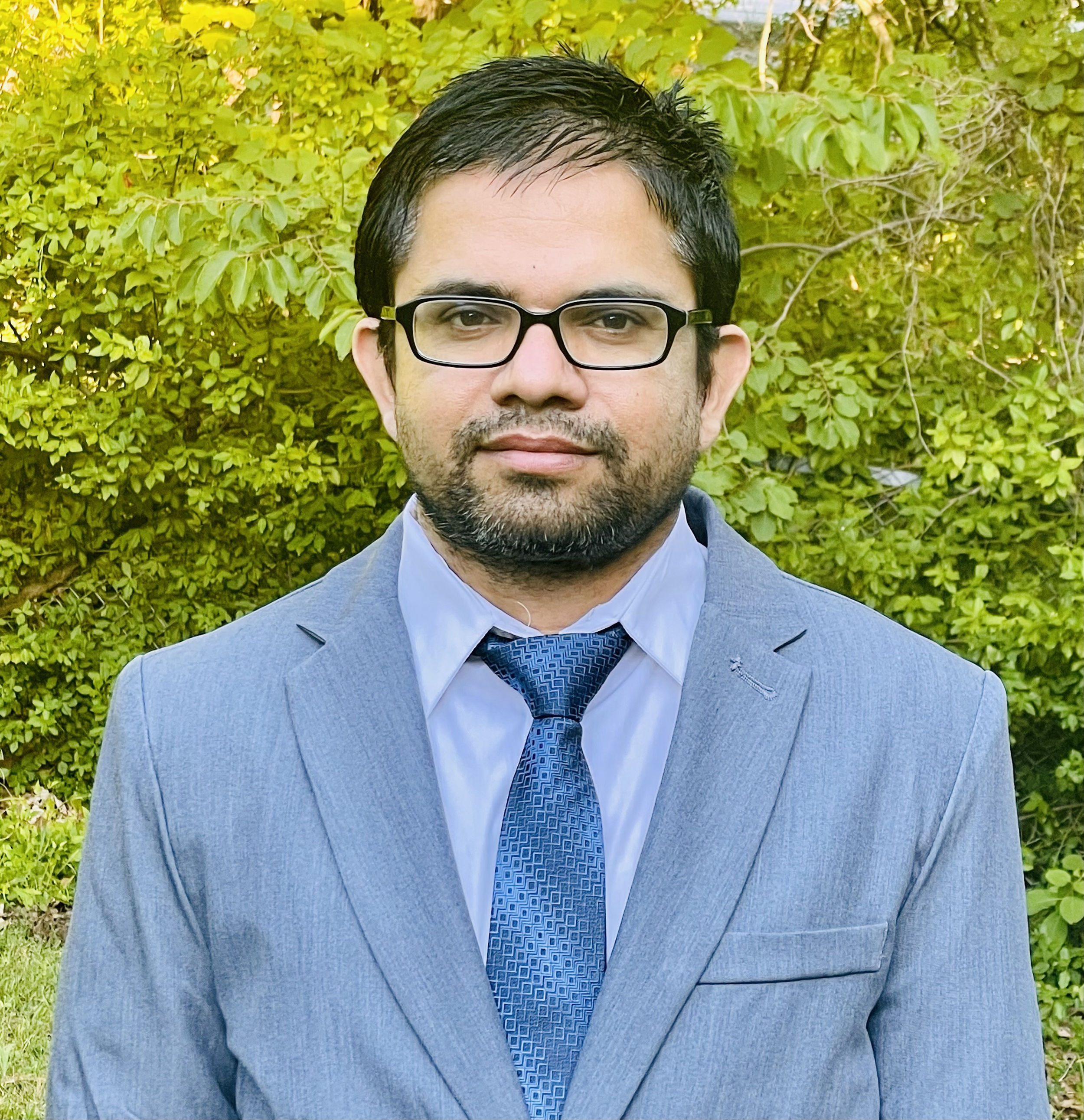 A man wearing a light blue suit, white shirt, and patterned blue tie, standing outdoors in front of green foliage.