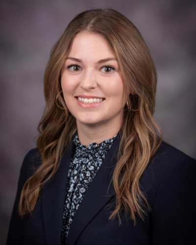 Professional headshot of a young woman with long, wavy brown hair, smiling, wearing a dark blazer and a patterned blouse against a gray background.
