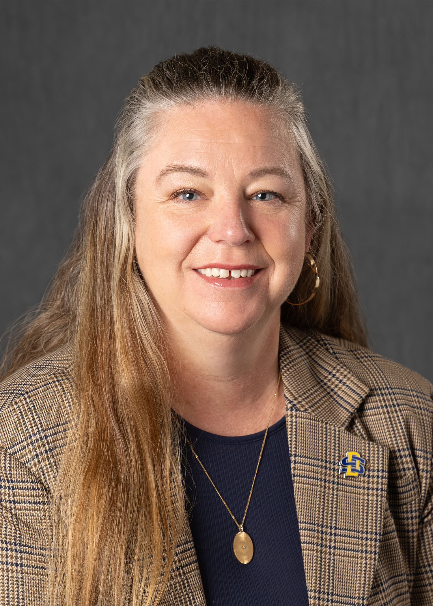 A woman with long, light brown hair, light skin, and wearing makeup, dressed in a plaid blazer over a navy top, gold necklace, and earrings, smiling against a dark gray background.