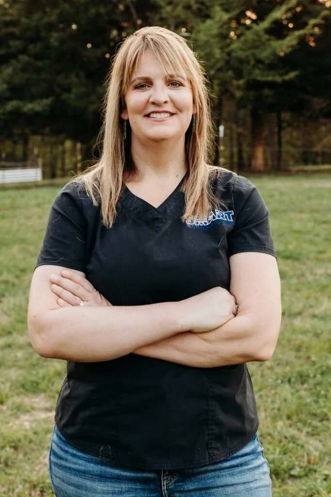 A woman with blonde hair and a black shirt standing outdoors with her arms crossed, smiling at the camera, in a grassy area with trees in the background.