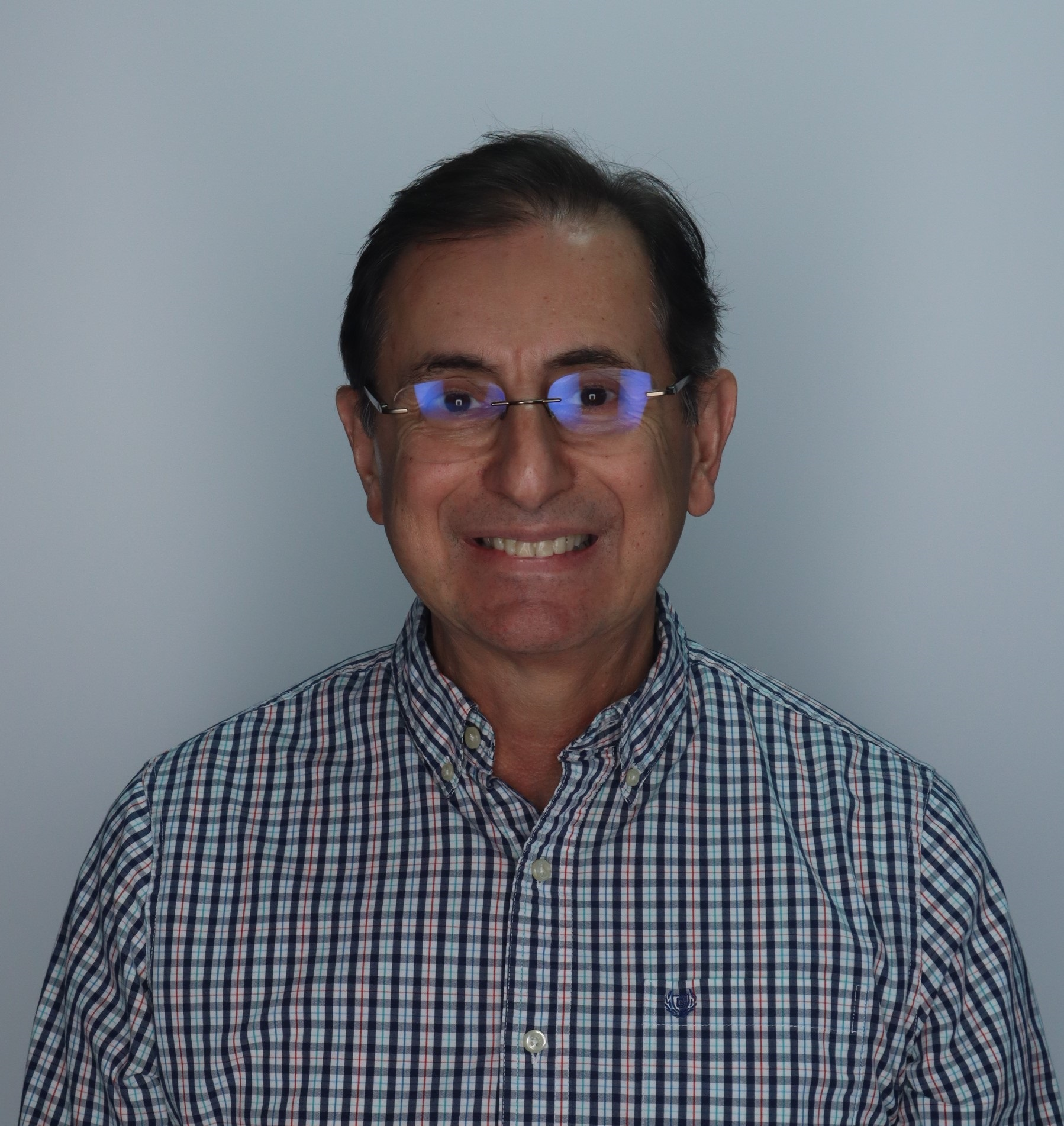 Portrait of a middle-aged man with dark hair, wearing glasses with blue reflections, and a plaid button-down shirt, smiling against a plain light gray background.