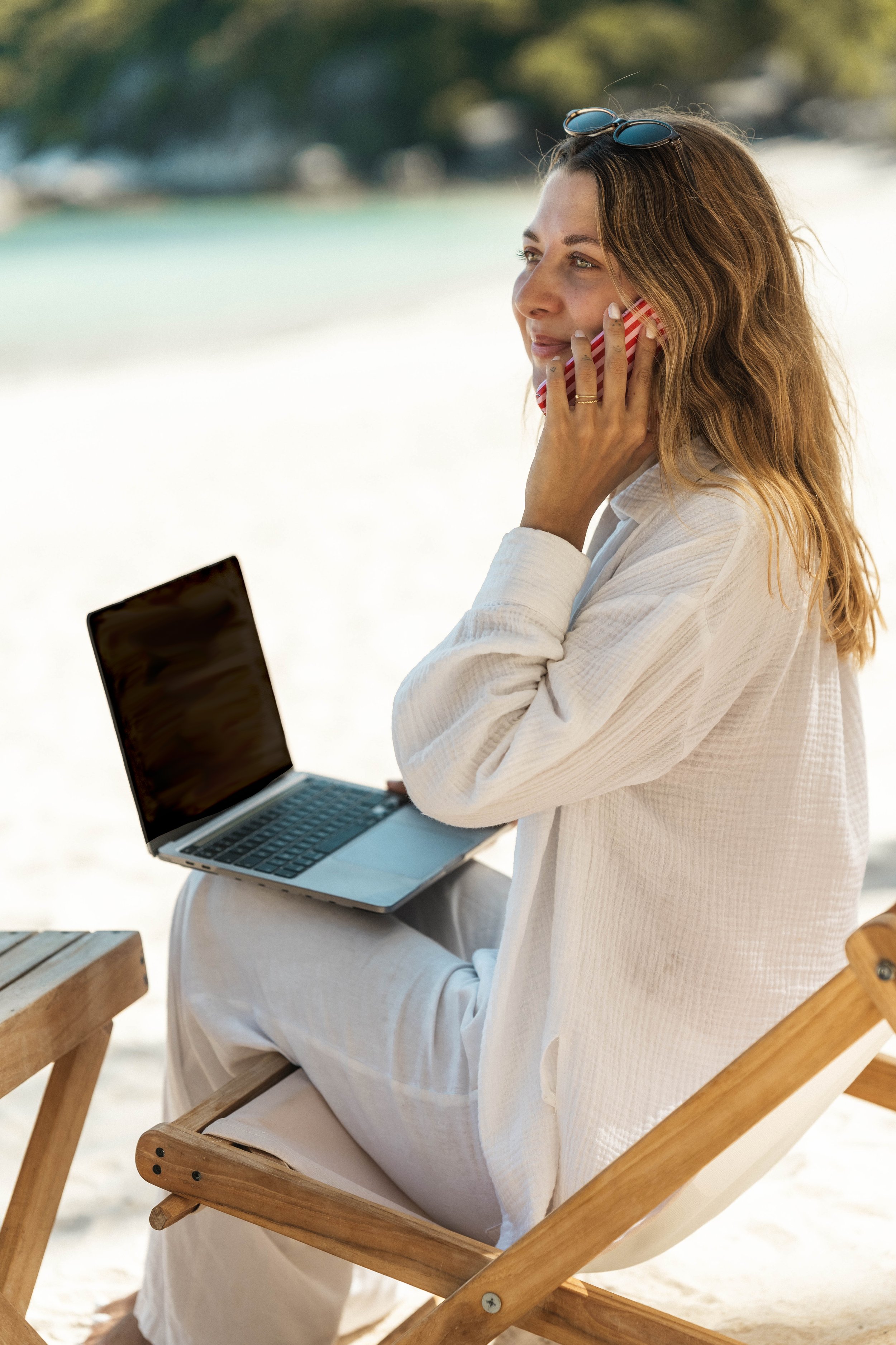 Een vrouw zit op een houten strandstoel, draagt een witte jurk en gebruikt een laptop en een telefoon. Ze heeft een zonnebril op haar hoofd en kijkt naar links.