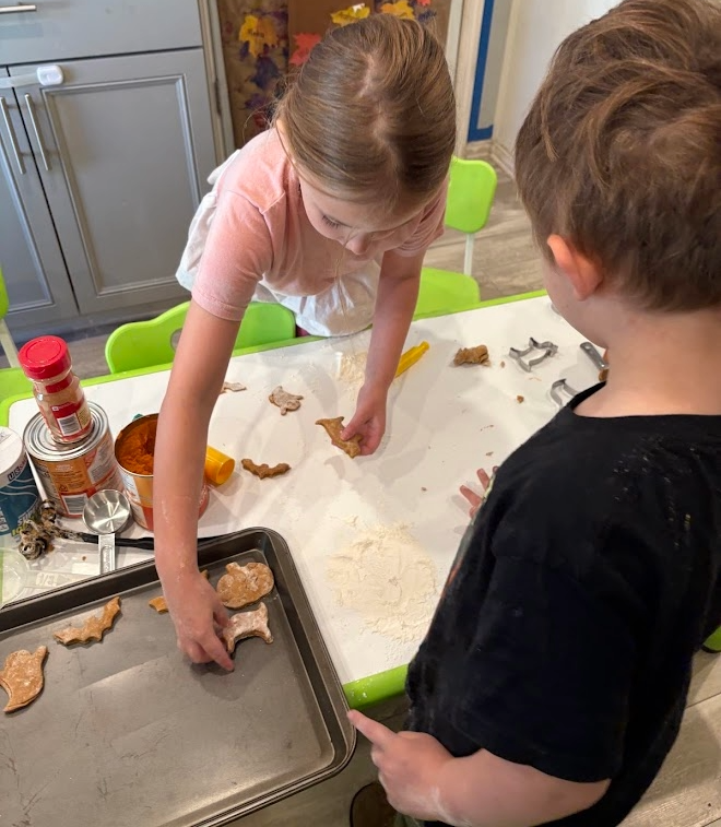 Two children baking cookies in a kitchen, with one girl placing cookie cutters on dough and a boy watching.