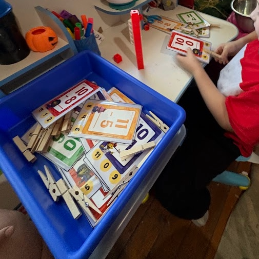 Child sitting at a table playing with number and math flashcards, with clothespins and colored markers nearby, on a cluttered table.