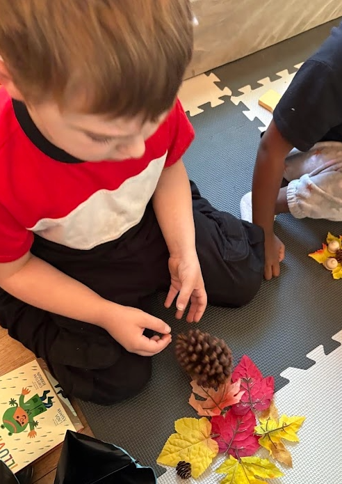 Child sitting on a foam puzzle mat, looking down at a nature craft project with pine cones and autumn leaves.